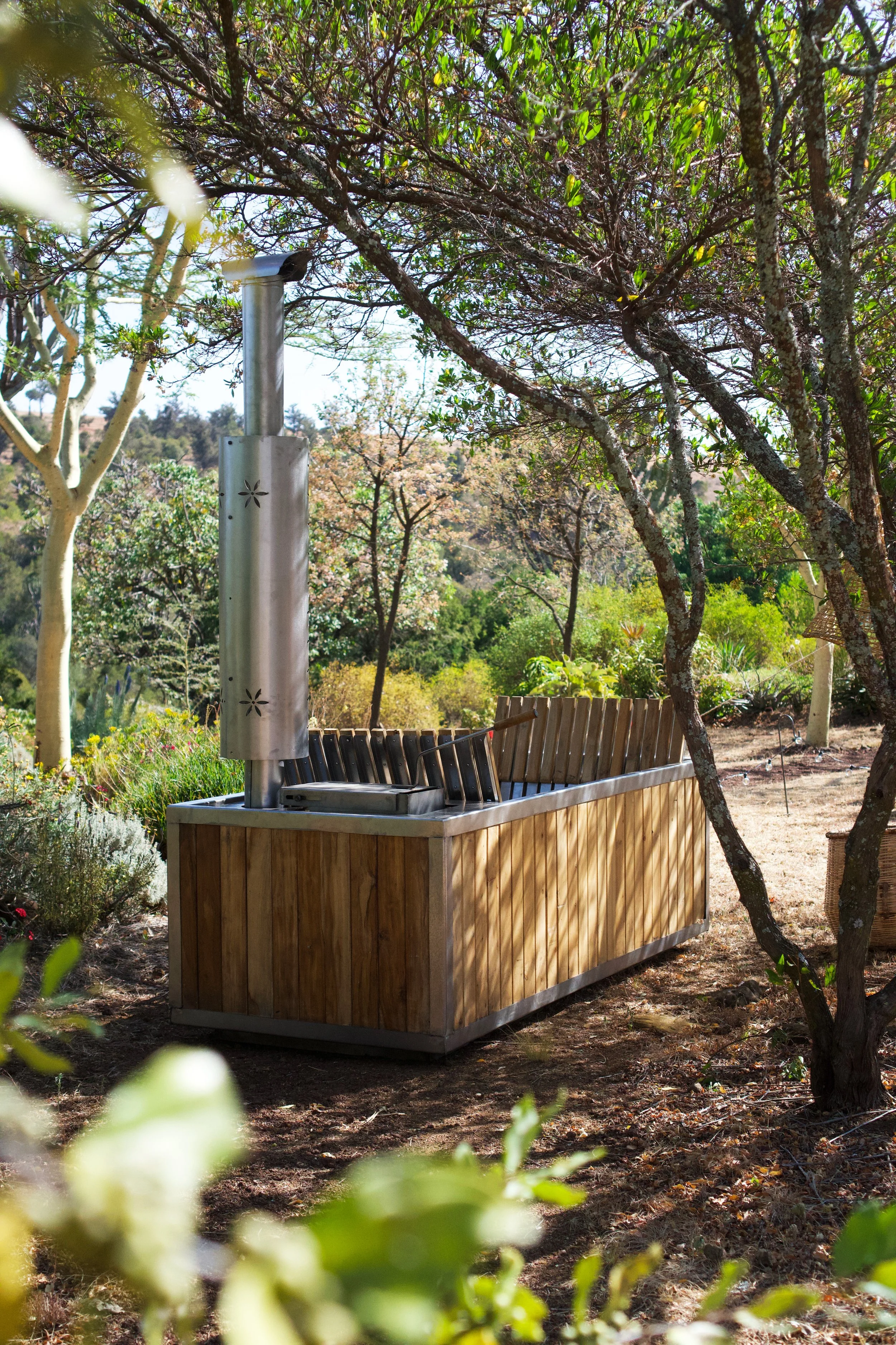 An outdoor scene featuring a wooden bar with a metal chimney, surrounded by trees, plants, and greenery on a sunny day.