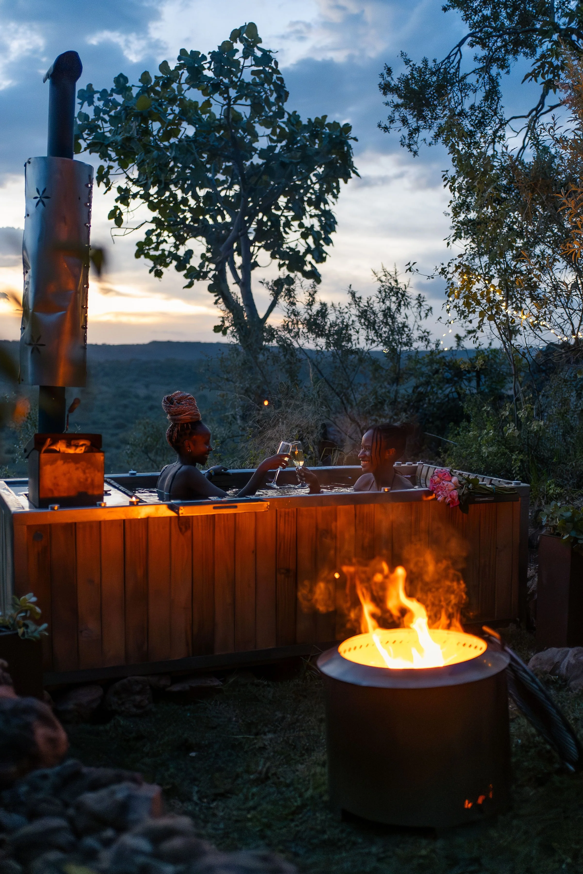 Two women relaxing in a hot tub outdoors during early evening with a fire pit in the foreground, surrounded by trees and hills, under a cloudy sky.