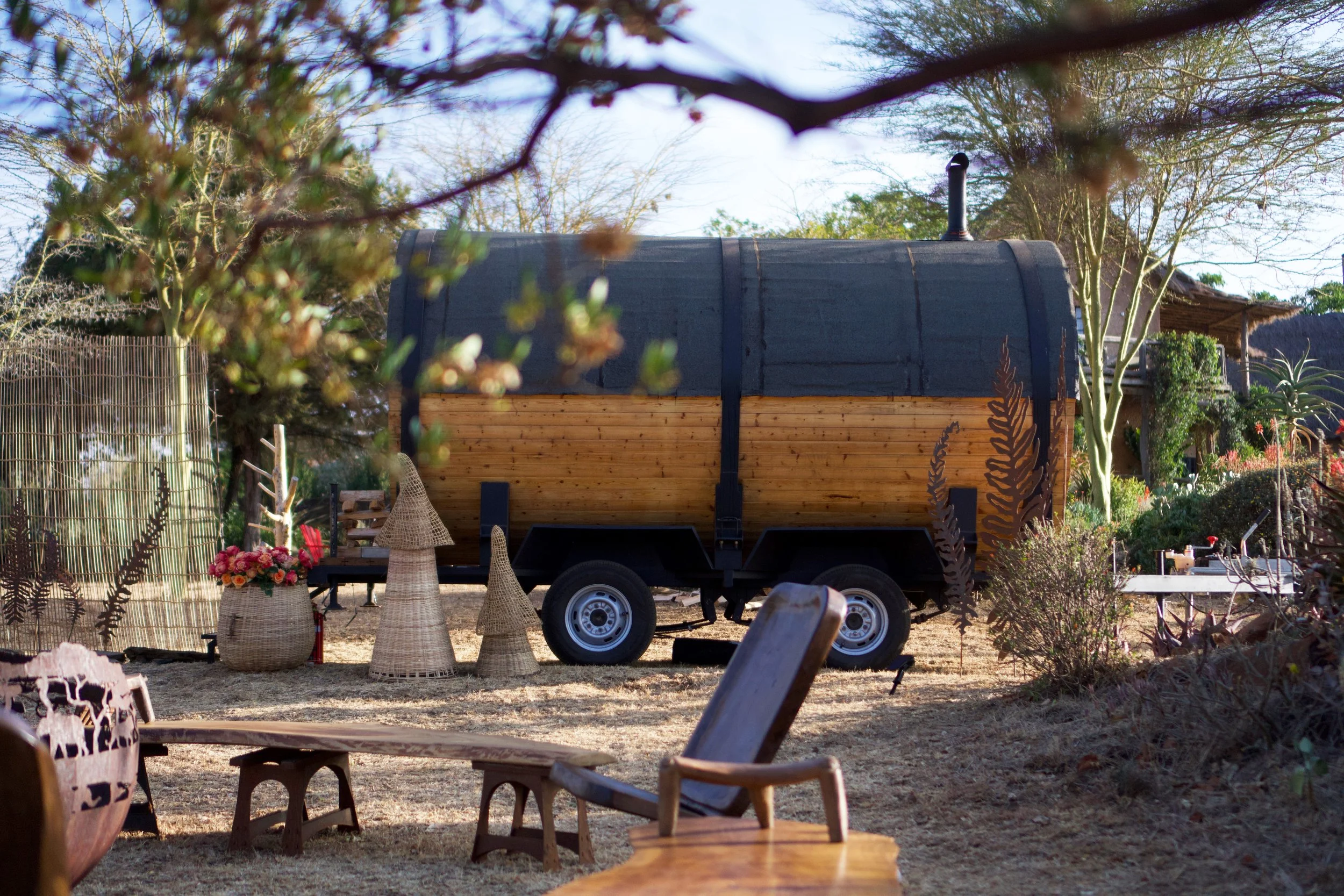 A rustic outdoor scene featuring a wooden trailer with a rounded roof, surrounded by trees and plants. There are wicker baskets with flowers and a variety of chairs in the foreground.