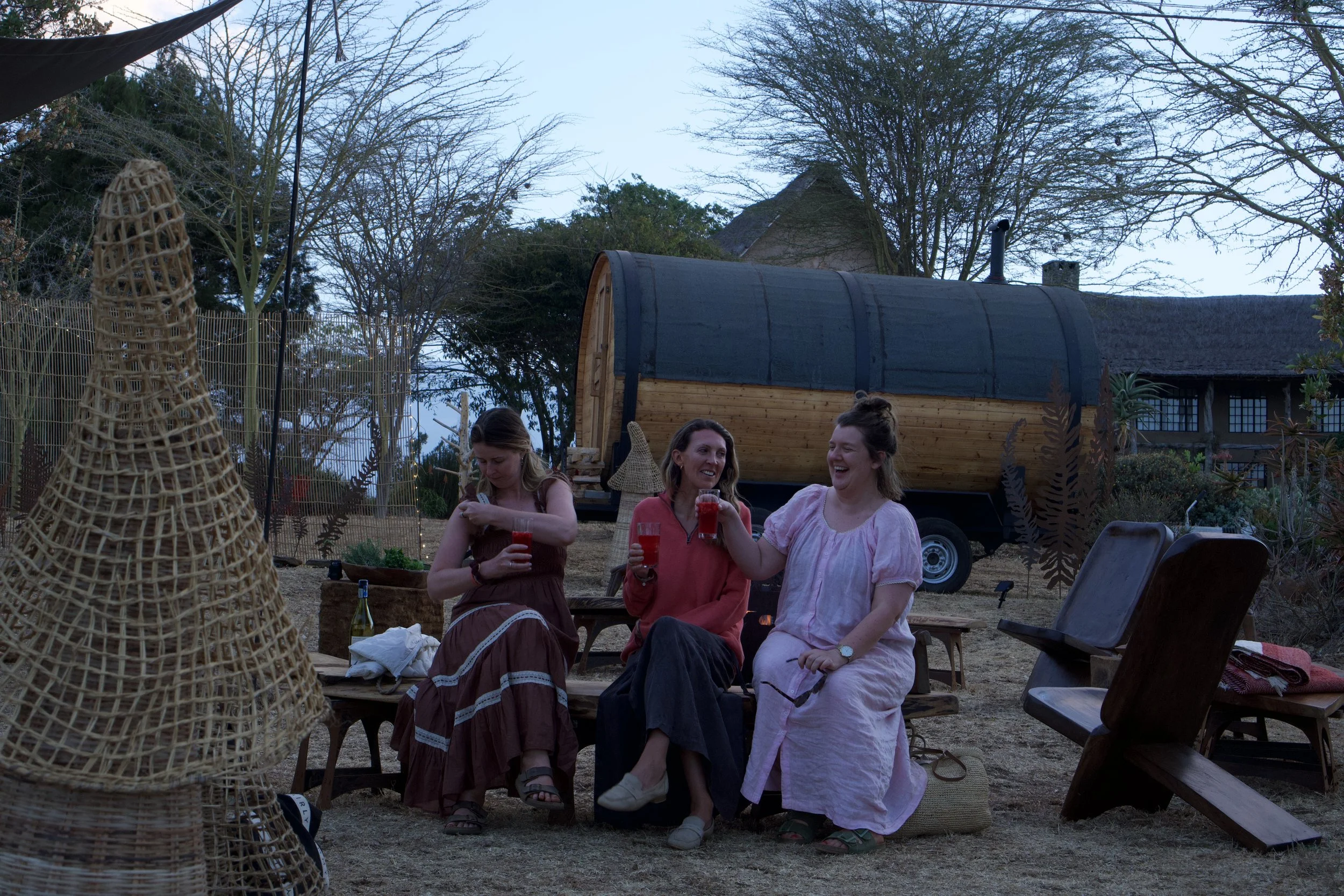 Three women laughing and holding drinks at an outdoor gathering during twilight, with a small trailer, trees, and a house in the background.