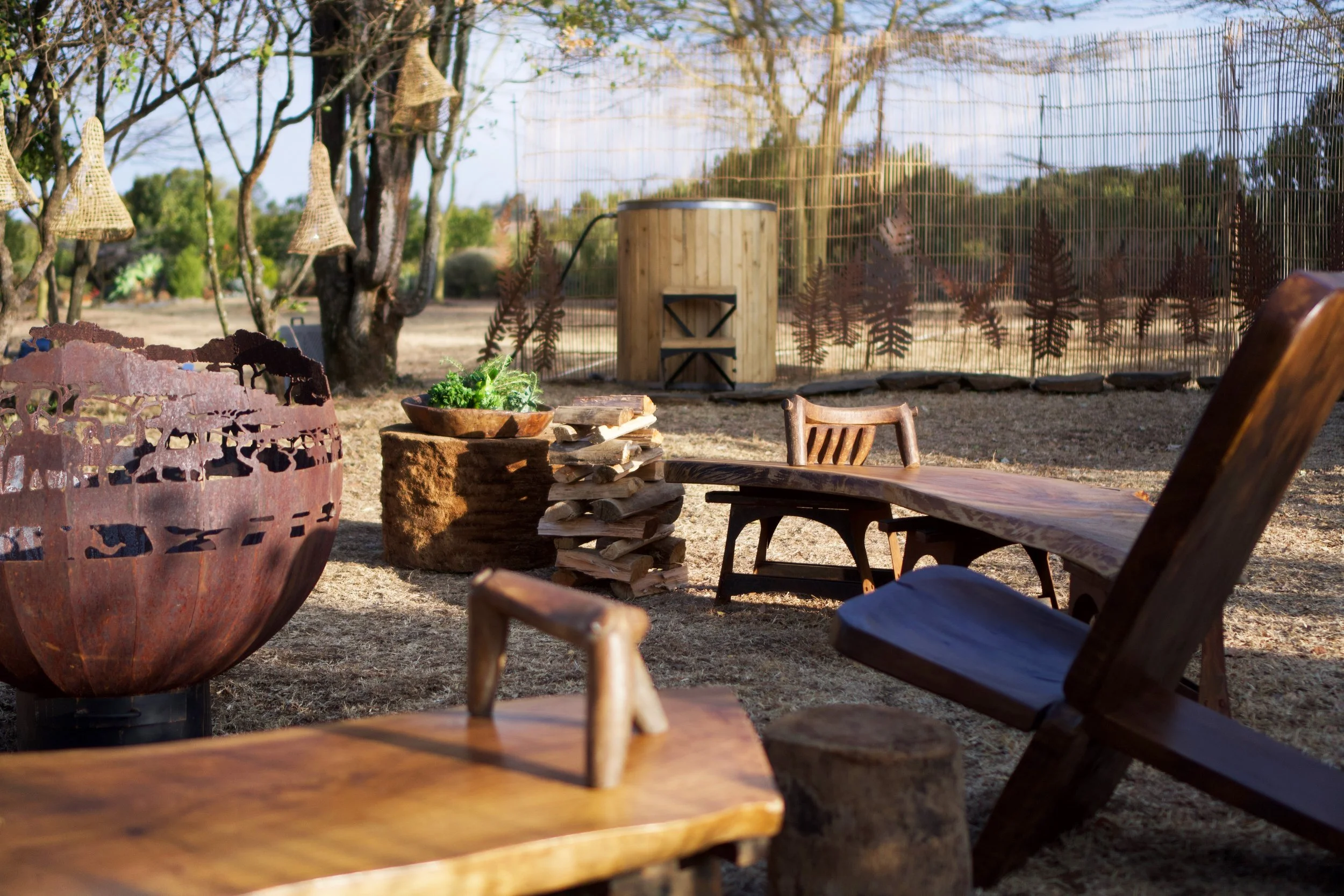 Outdoor seating area with wooden chairs and a large metal fire bowl, trees, and a bamboo fence in the background.