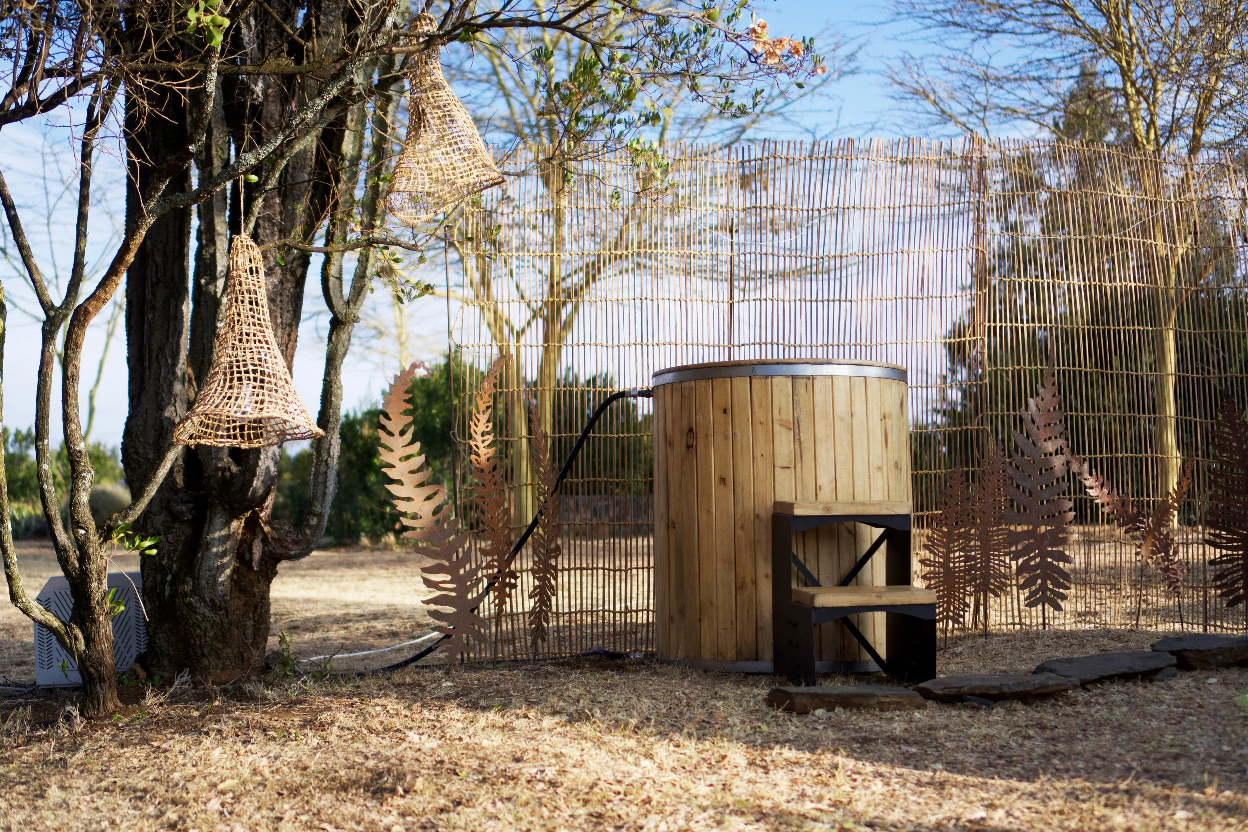A garden scene with a tree on the left and a bamboo privacy screen in the background. Hanging from the tree are woven mesh feeder bags, and there is a wooden cylindrical structure, possibly a planter or compost bin, beside the screen. There are some 