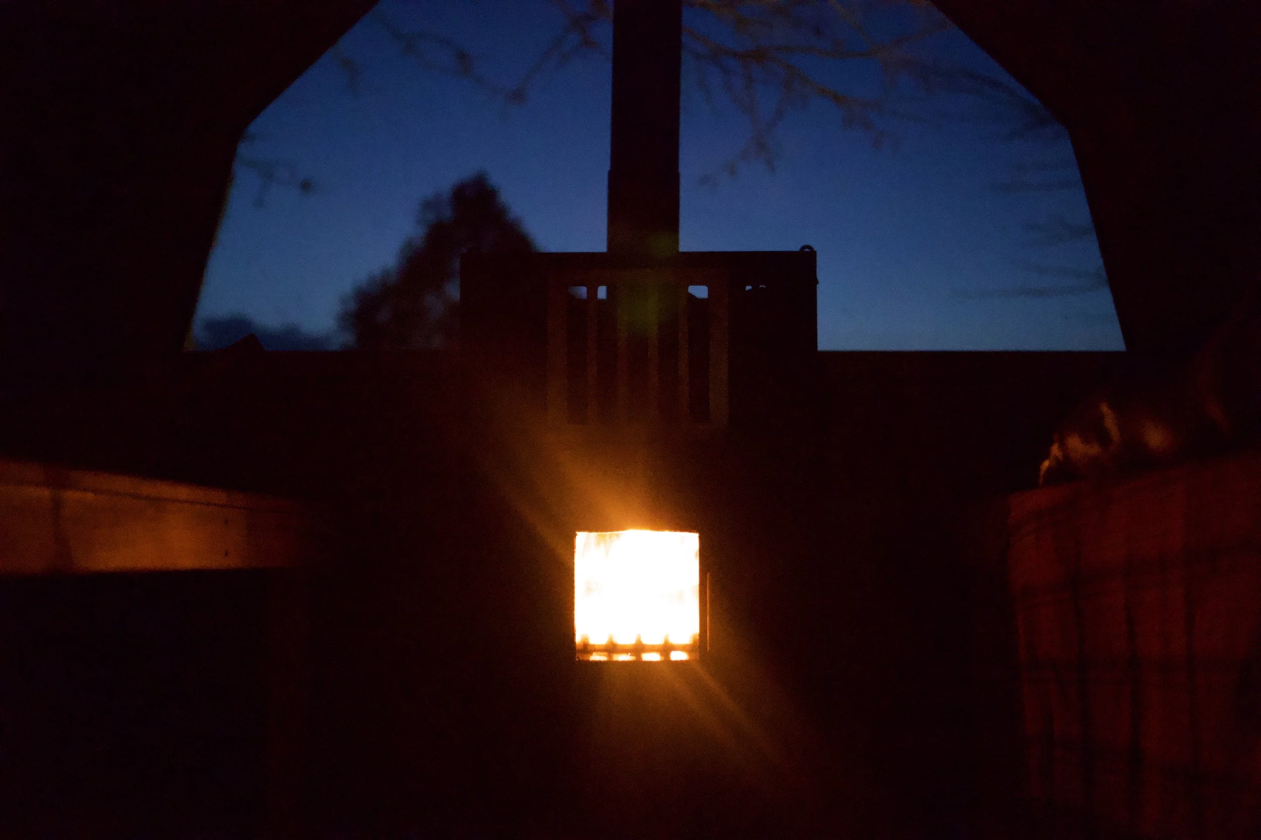 Photo taken from inside a tent during dusk showing a silhouetted tree and chimney in the background with a glowing light inside the tent.