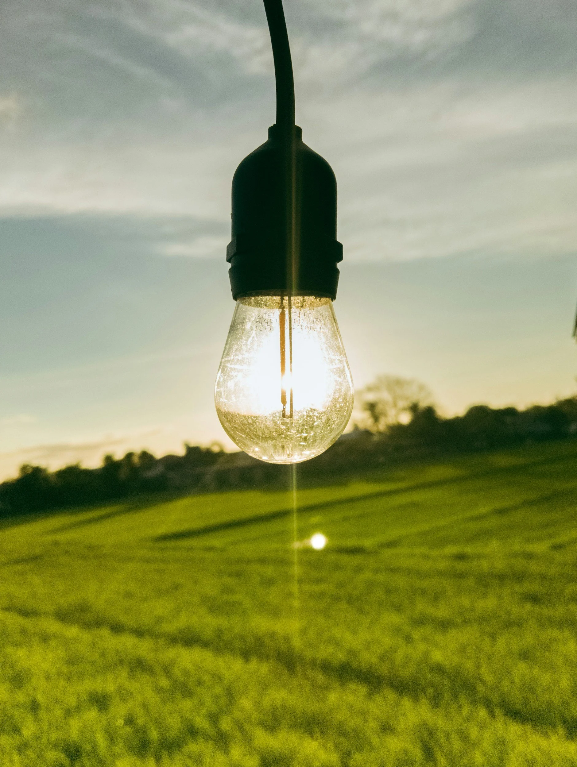 Close-up of a hanging light bulb against a cloudy sky at sunset with a grassy field below.