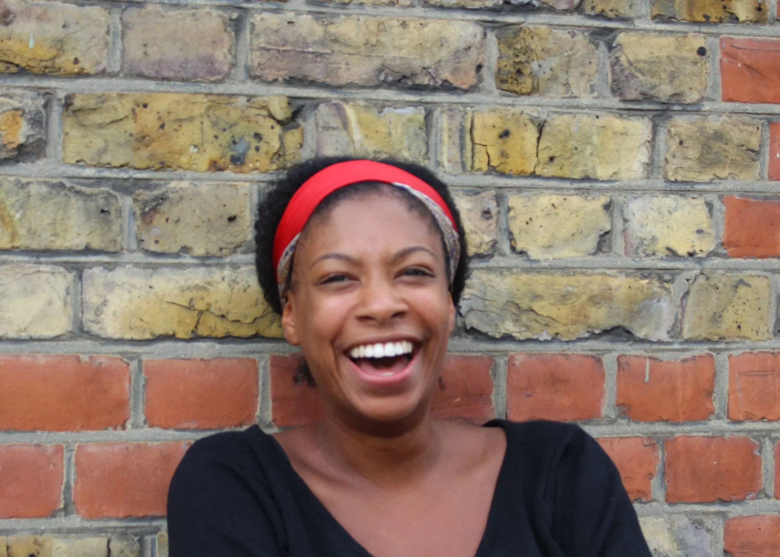 A woman smiling and laughing in front of a brick wall, wearing a red headband and a black shirt.