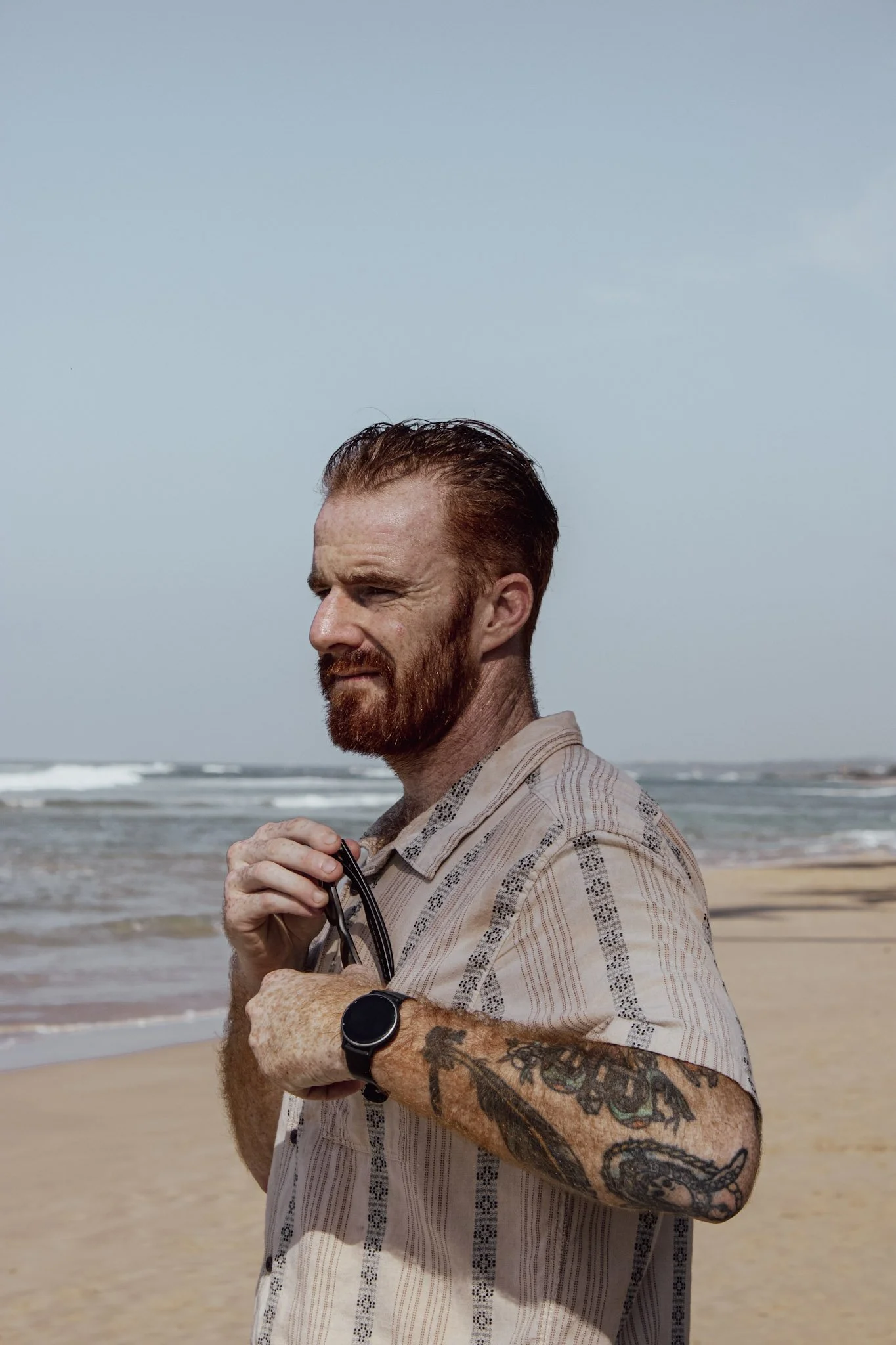Cam Springett on a beach, holding his sunglasses and looking out along the shore