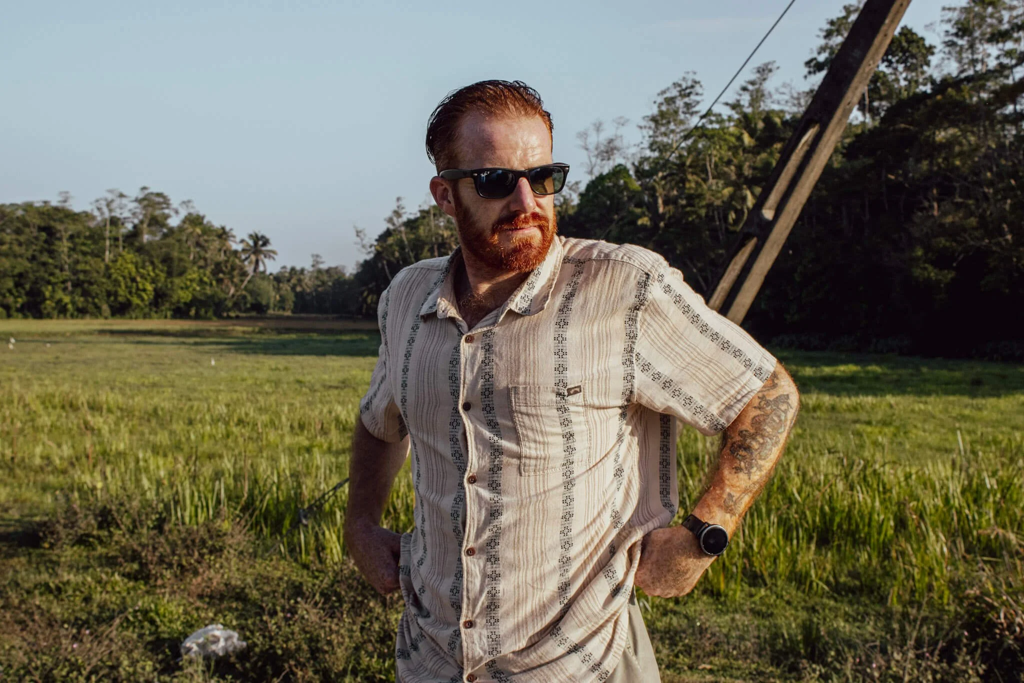 Cam Springett, online sobriety and performance coach, standing at the edge of a paddy field in warm evening light in Ahangama, Sri Lanka, wearing sunglasses and a linen shirt.