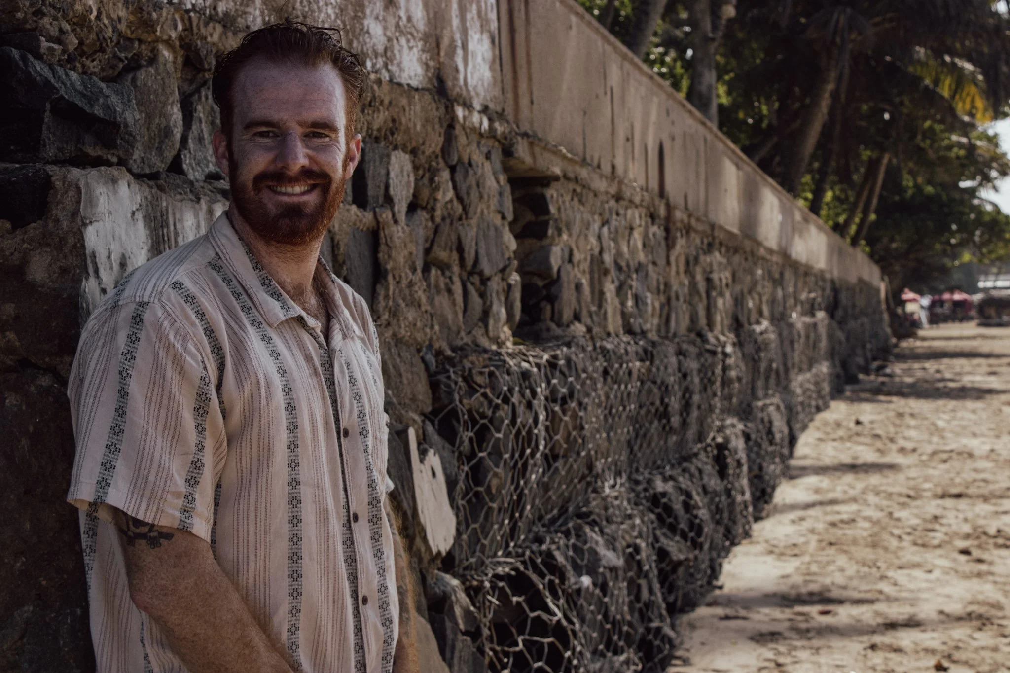 Cam Springett smiling beside a stone sea wall lined with palm trees on a beach in Sri Lanka