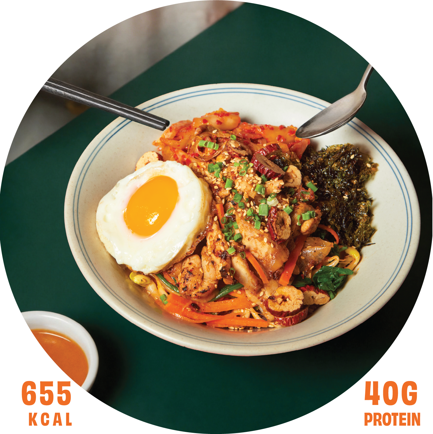 A bowl of Asian-inspired mixed dish with noodles, vegetables, chopped green onions, sesame seeds, and sliced meat, topped with a fried egg, accompanied by a small bowl of sauce.