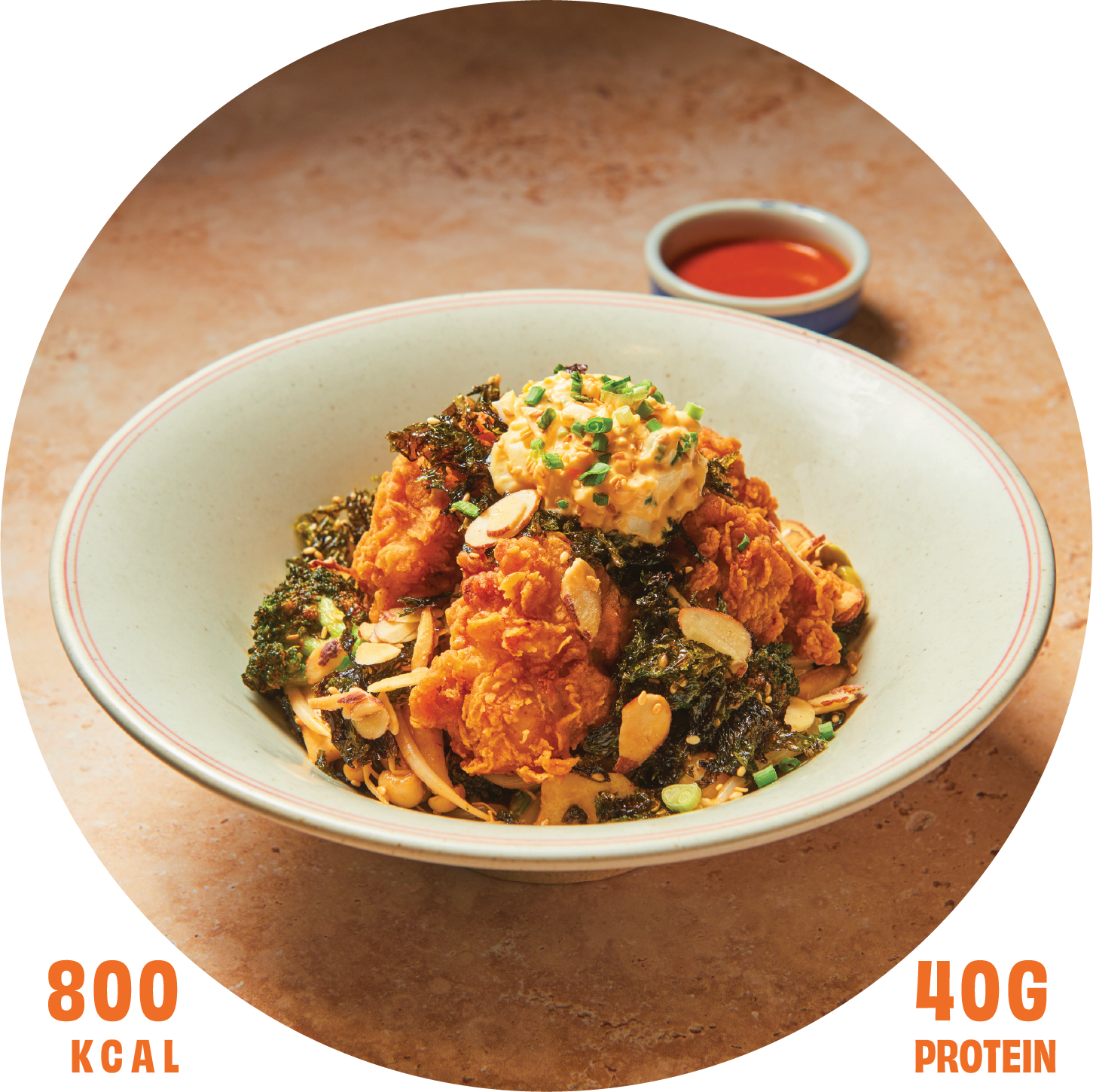 A bowl of Asian fried chicken topped with a scoop of potato salad, chopped green onions, and sliced almonds, with a small dish of red dipping sauce in the background.