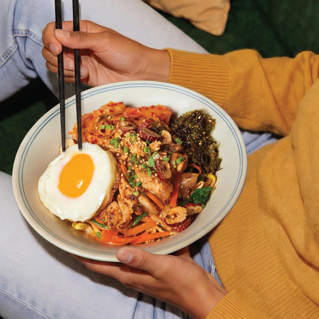 A person wearing a yellow sweater holding a bowl of Korean bibimbap topped with a fried egg, vegetables, and sesame seeds, using chopsticks.