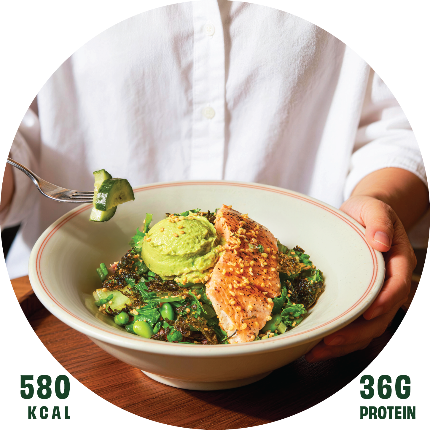 A person in a white shirt holding a bowl with grilled salmon, mashed avocado, green peas, and leafy greens, garnished with seeds and herbs.
