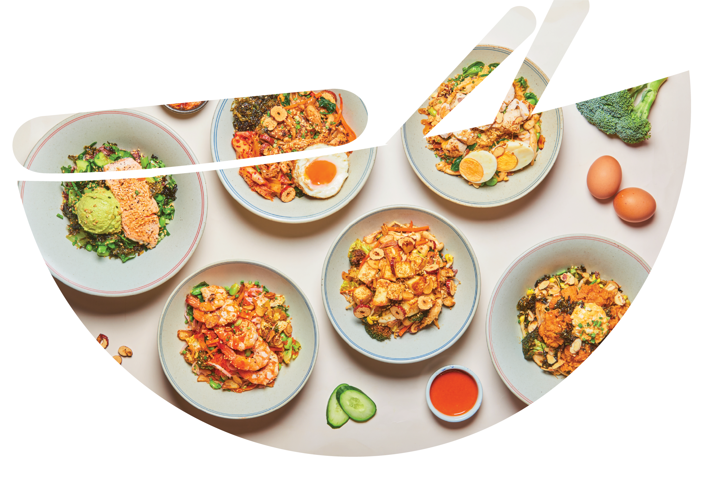 A variety of Asian-style dishes in bowls on a white table, with cucumber slices, broccoli, eggs, and small bowls of sauce nearby.
