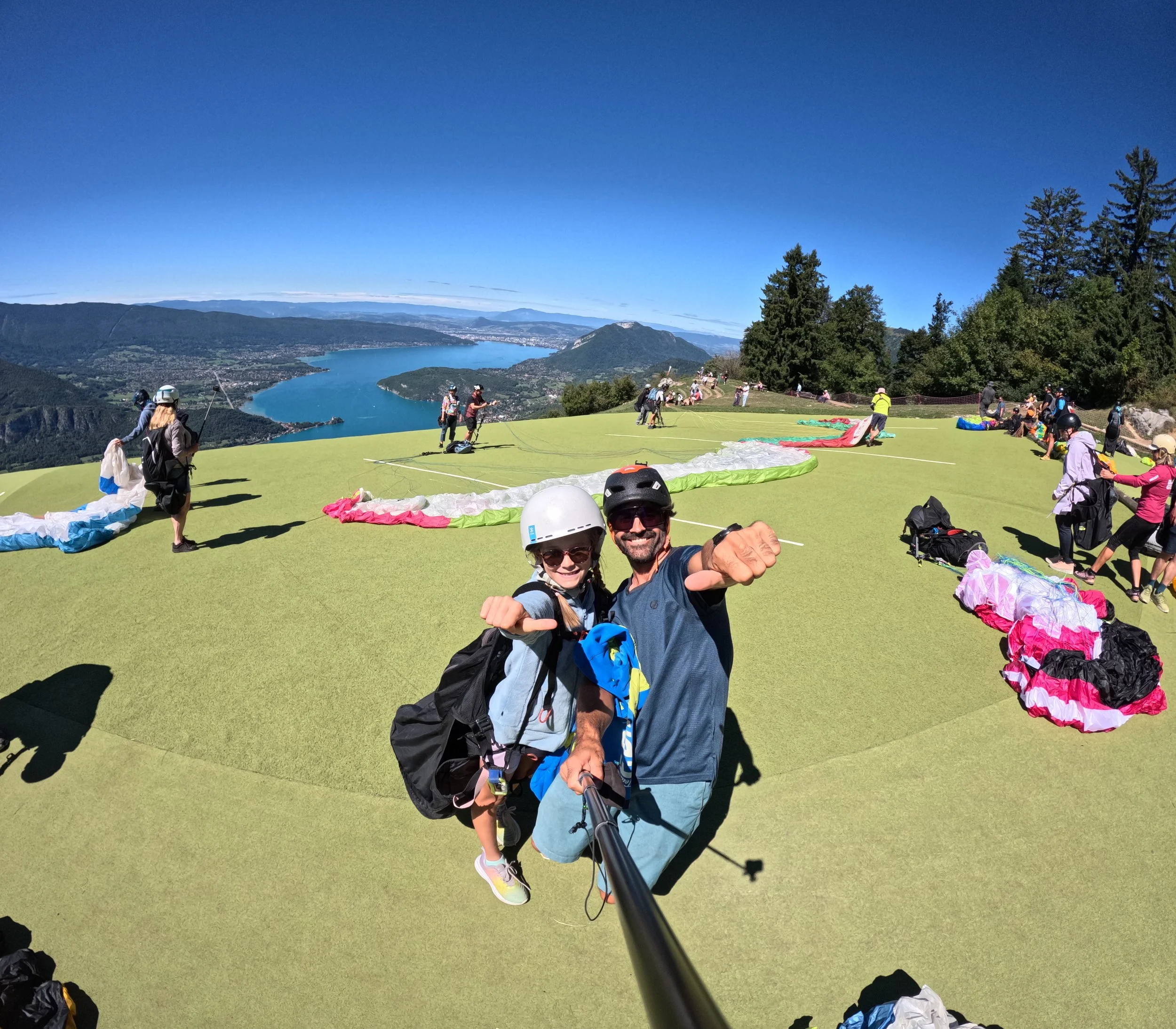 Deux personnes portant un casque de protection prennent une selfie sur une colline vert vif avec des parapentes déployés en arrière-plan et un lac entouré de montagnes dans le paysage.