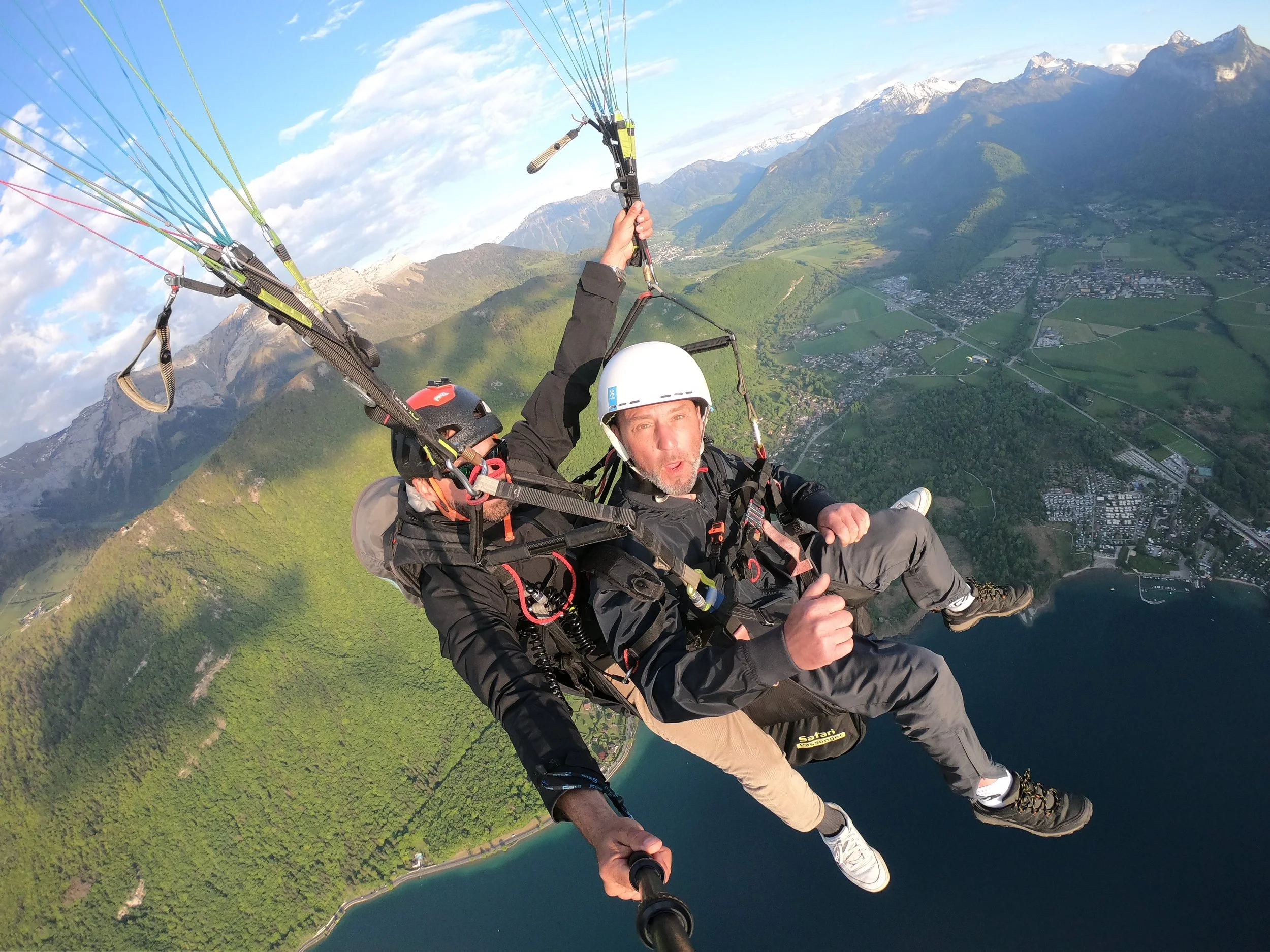 Deux personnes en tandem de parapente volant au-dessus d'une vallée avec montagnes et un lac, portant des casques de sécurité.