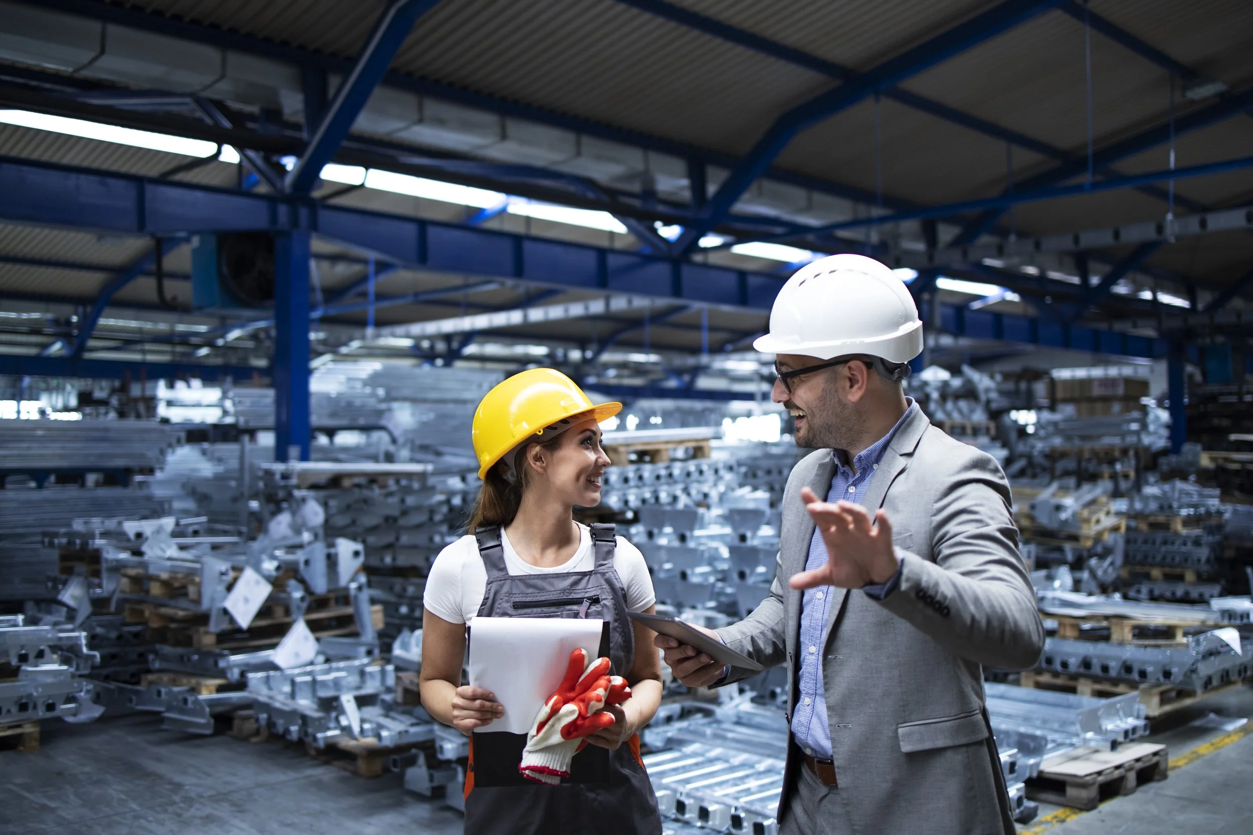 Two professionals, a woman and a man, wearing safety helmets in a warehouse, having a conversation. The woman holds a clipboard and a pair of gloves, and the man appears to be explaining or discussing something with her.