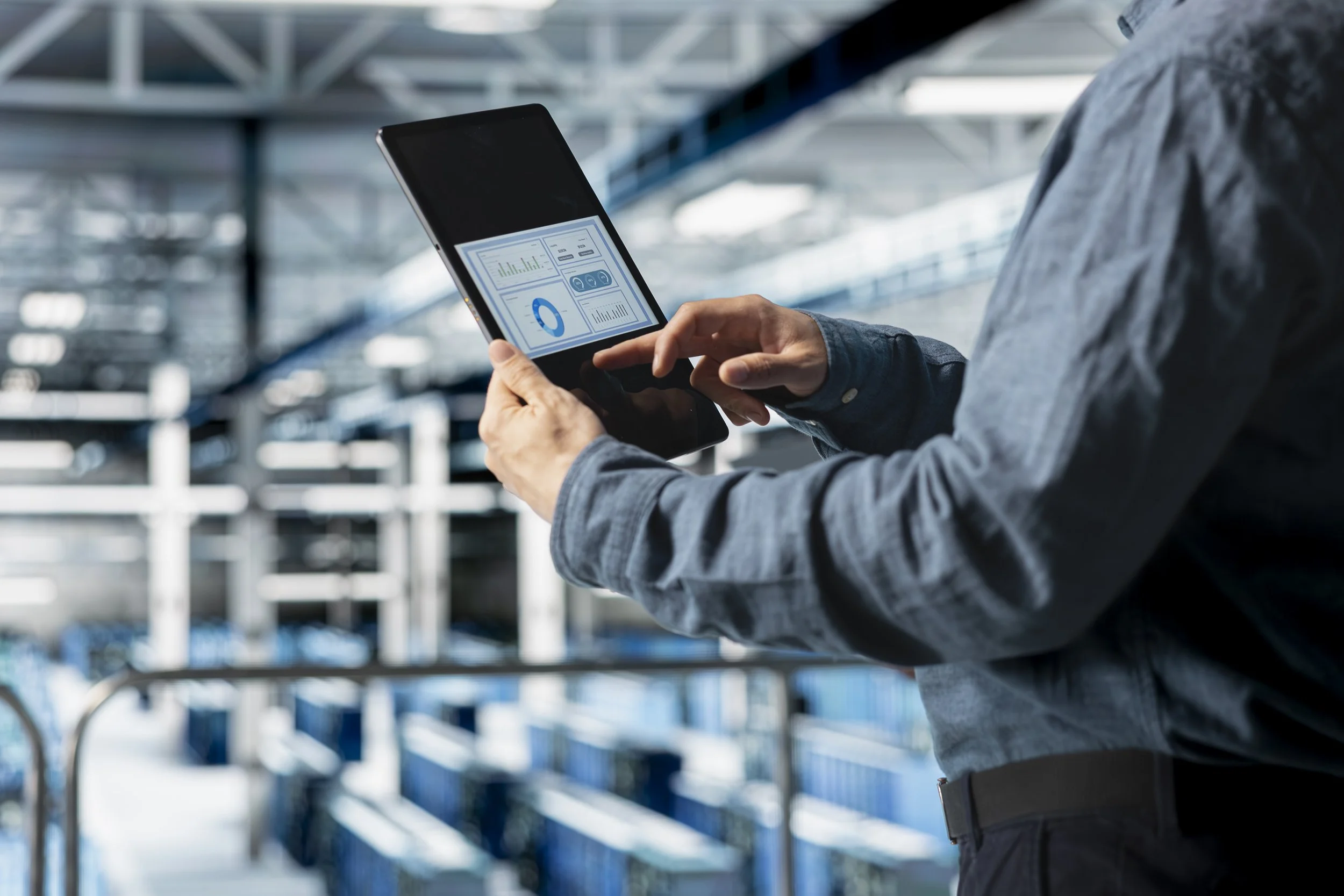Person in business attire using a tablet computer with graphs and charts displayed, in a data center or server room with metal shelves and servers.