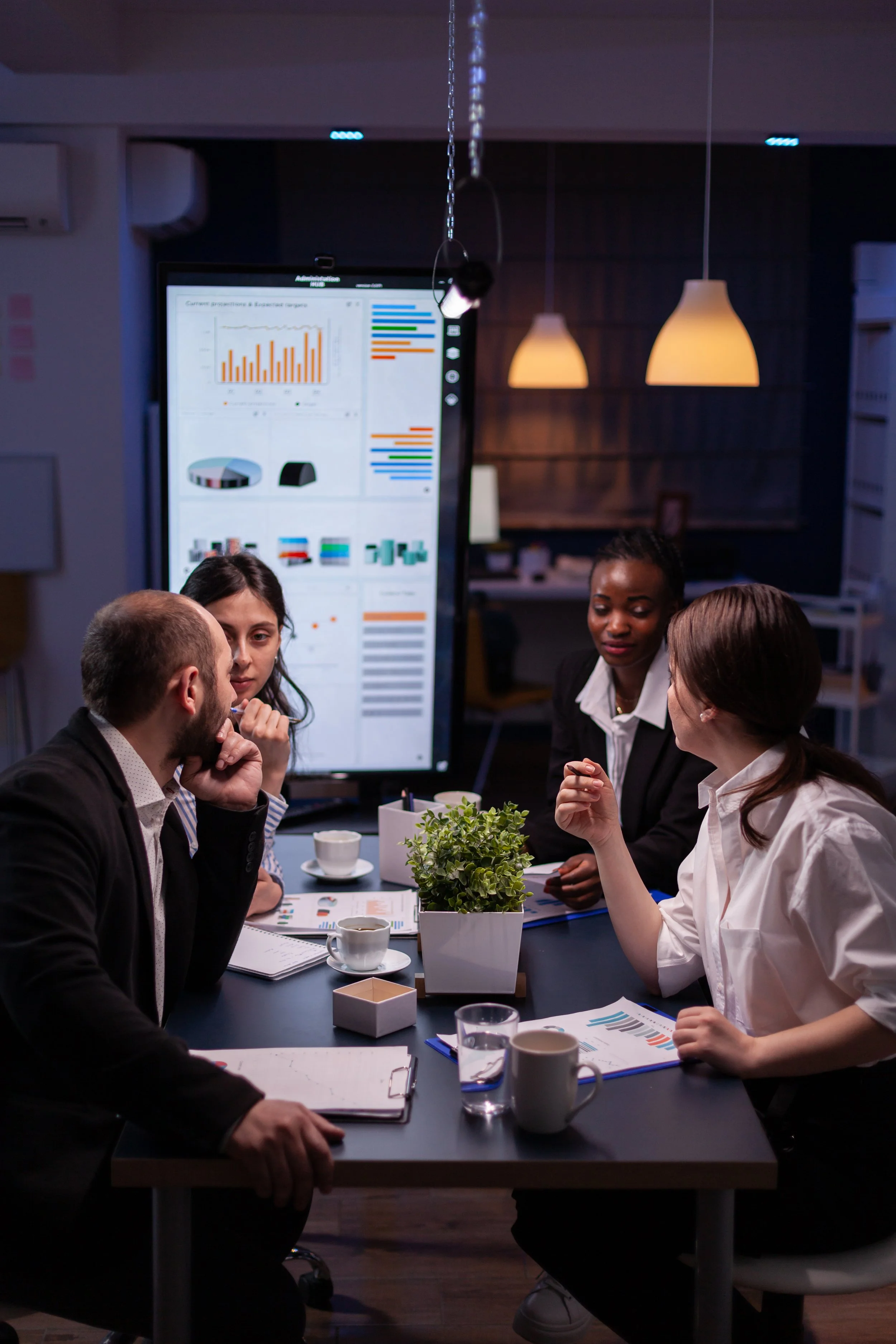 A group of five professionals seated around a table in a meeting room, engaged in discussion, with a large screen displaying charts and graphs in the background.