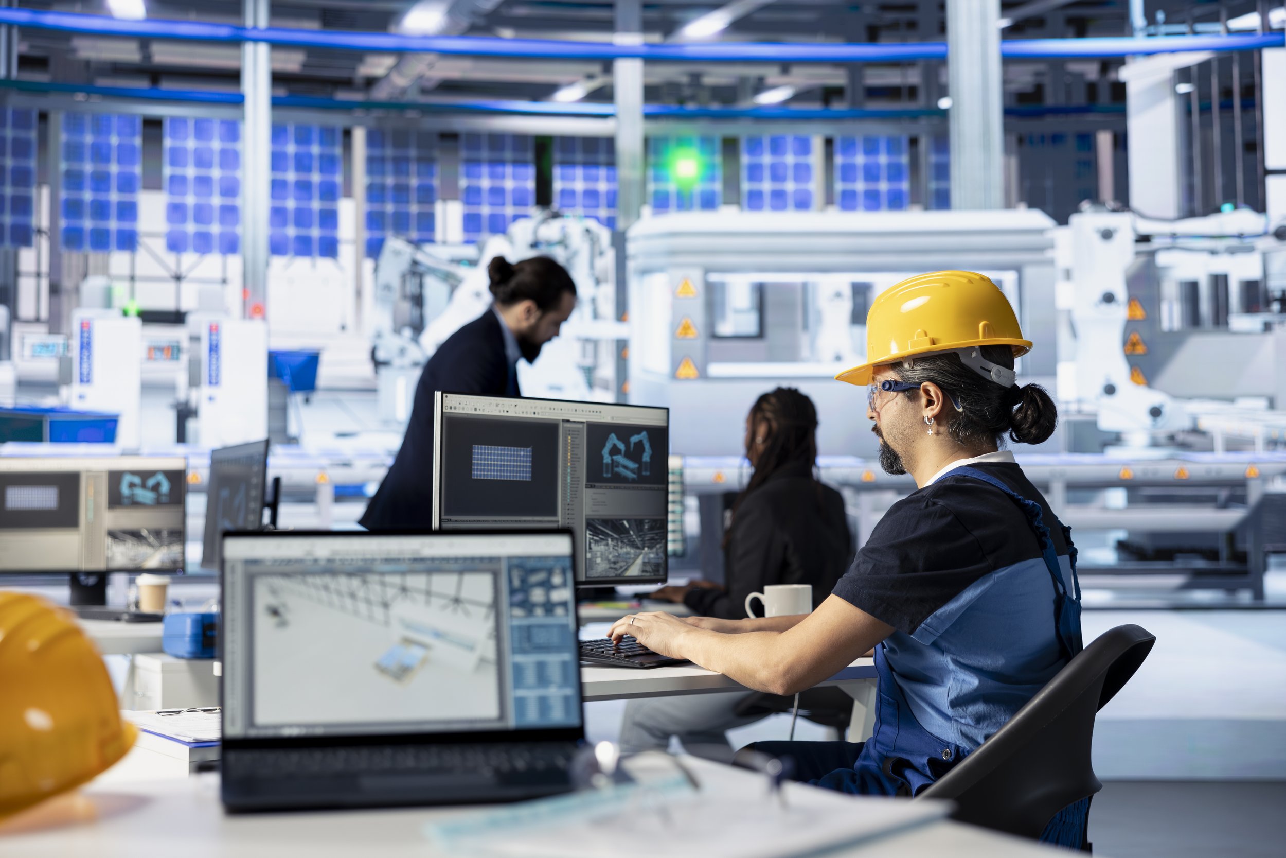A group of engineers working in a high-tech industrial or research facility with computer screens and advanced machinery, one wearing a yellow safety helmet.