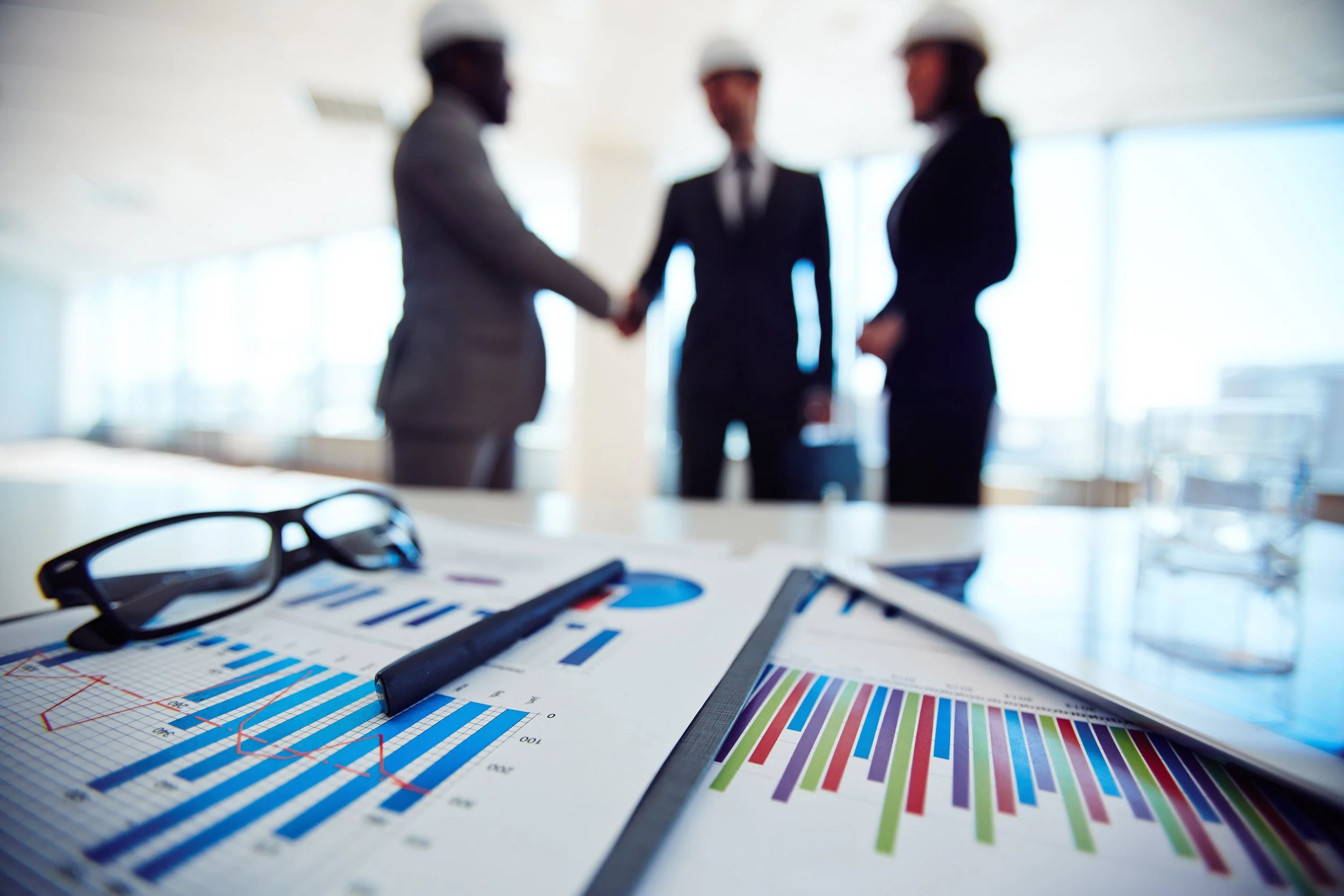Colorful charts, graphs, and documents on a desk with professional people wearing hard hats in a business meeting.