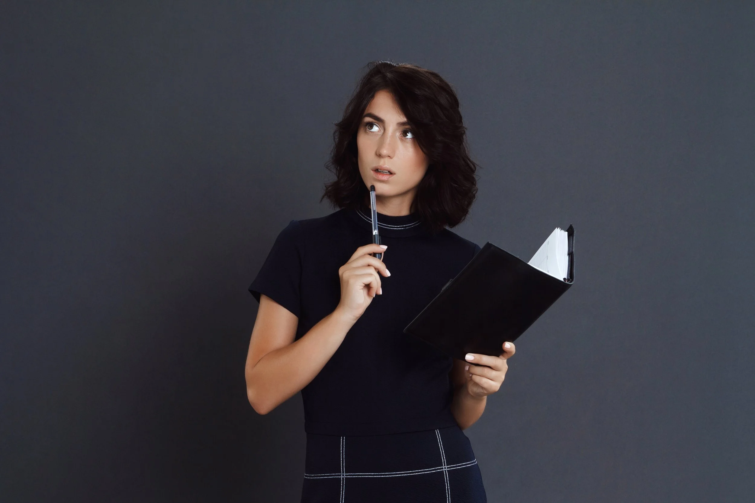 A young woman with dark, wavy hair, wearing a black top, is holding a black notebook and a pen, with her finger on the pen near her lips, looking thoughtfully to the side against a plain gray background.