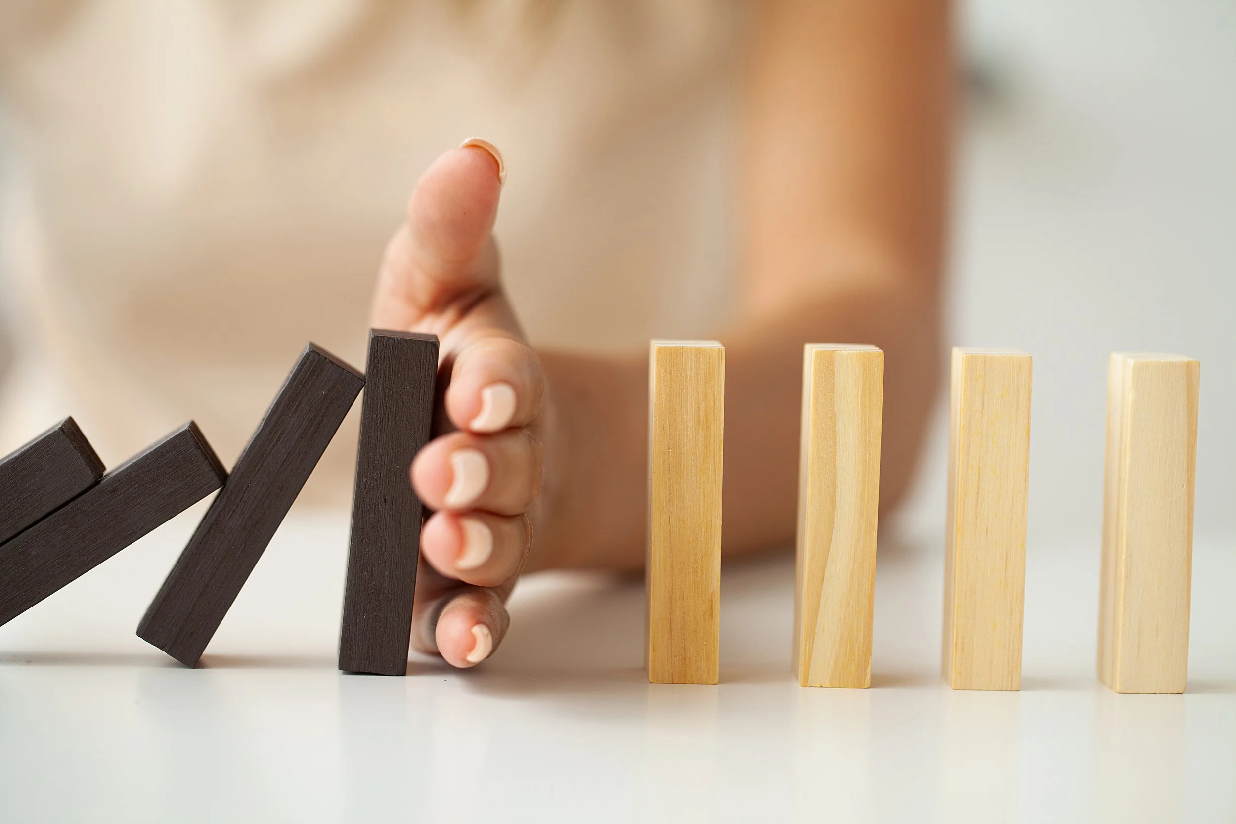 A hand stops black dominoes from falling, with natural wood-coloured dominoes standing upright on the right side.