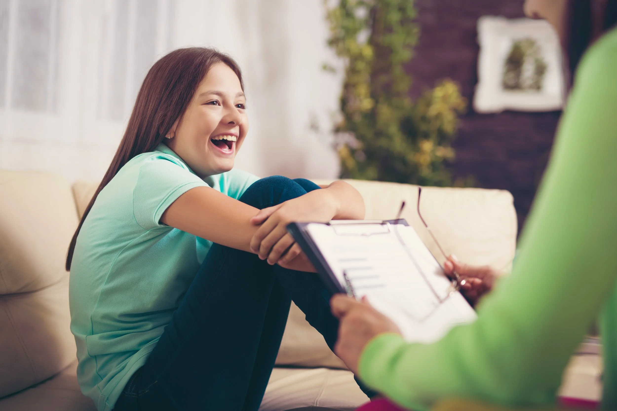 A girl with long brown hair, wearing a light blue shirt, laughing and sitting on a beige couch, talking to an adult who is holding a clipboard and glasses.