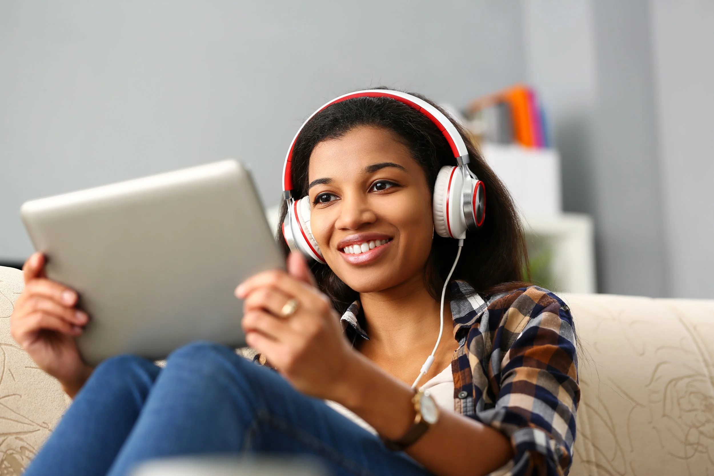 A young, BIPOC woman with dark hair wearing headphones and a plaid shirt sitting on a beige couch using a tablet. She is smiling and looks relaxed and comfortable.