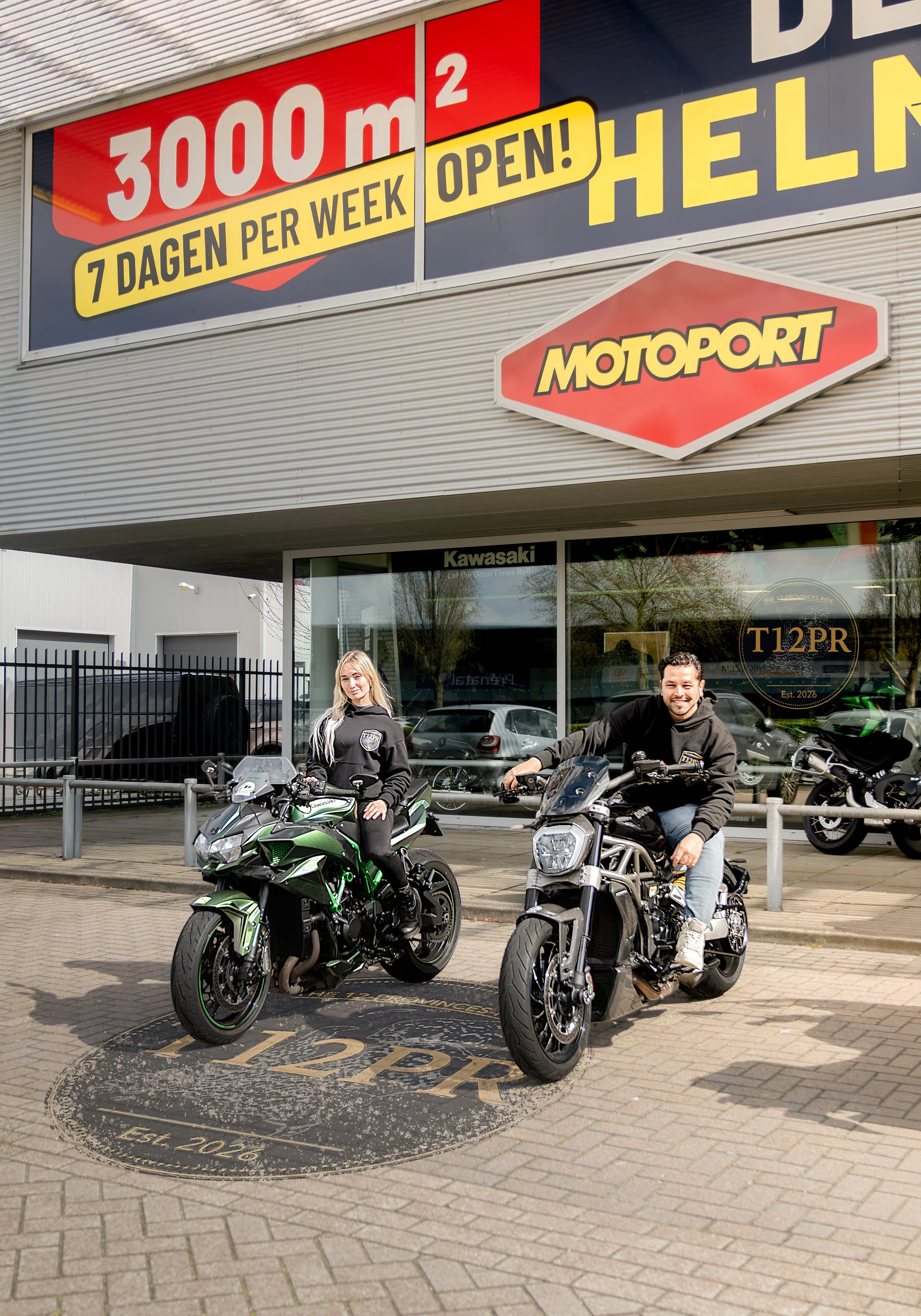Twee mensen op motoren voor een motorwinkel met grote posters en bedrijfsnaam in het raam, één man en één vrouw.