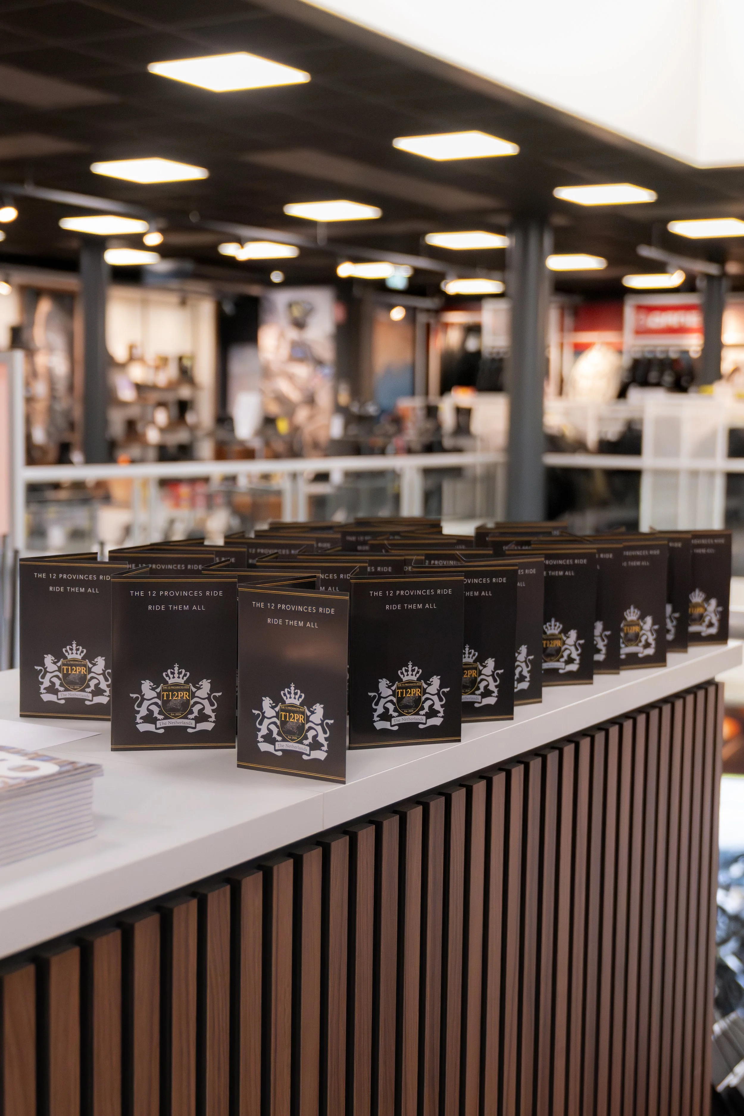 Kaarten met het logo en slogan van T12PR op een witte balie in een winkel of kantoor. In de achtergrond is een donkere interieurruimte met verlichting en diverse meubels.