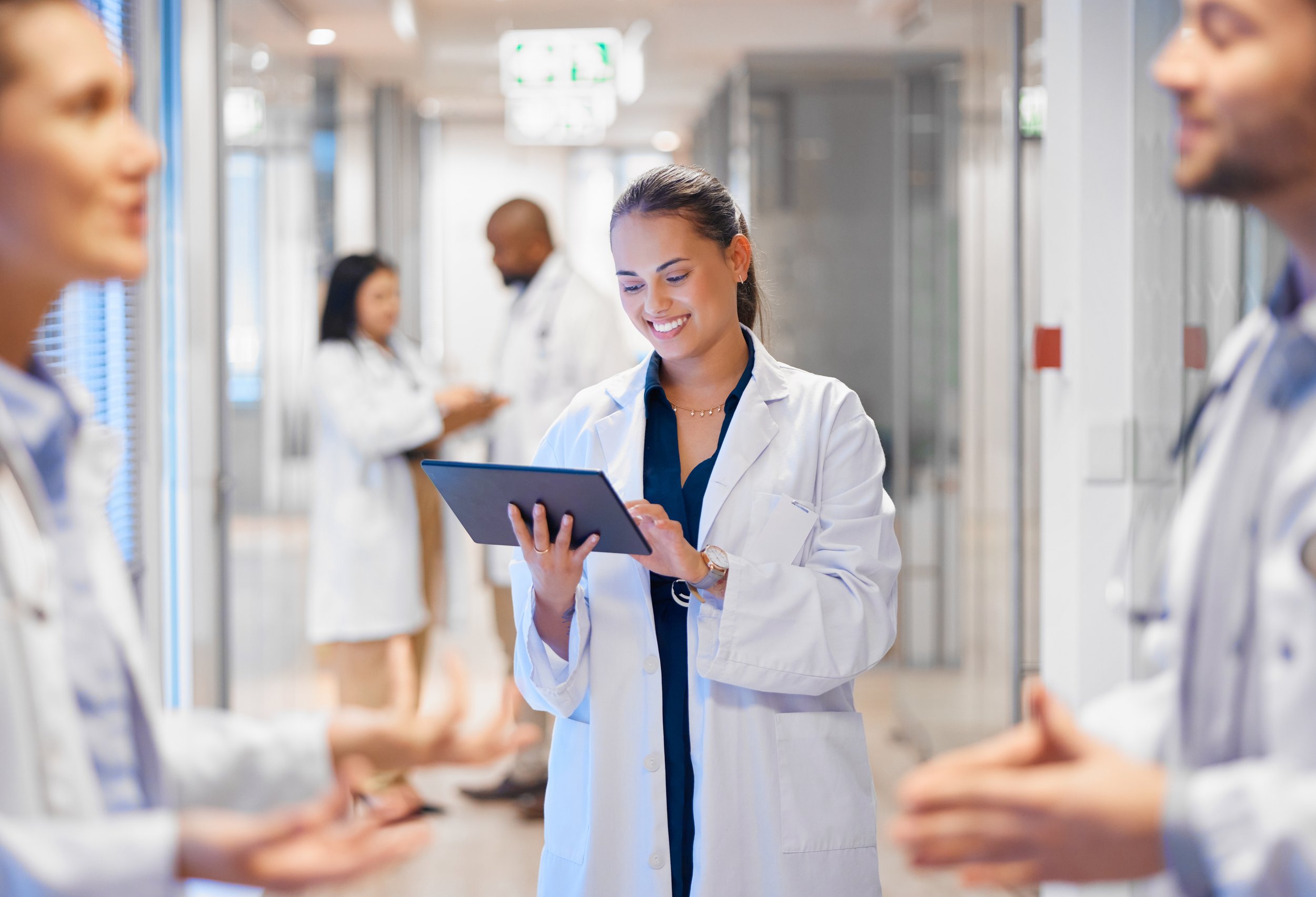 Eine Gruppe von Medizinern in einem Klinikflur, eine junge Frau in der Mitte liest auf einem Tablet, umgeben von Kollegen.