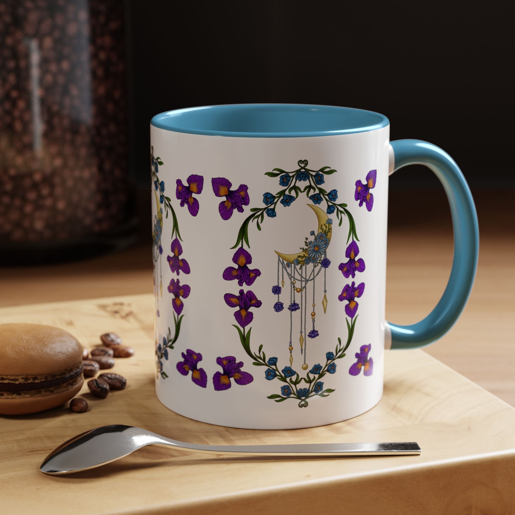 A ceramic mug with a floral design, a blue interior, and a blue handle on a wooden surface, with a spoon in front of it and some chocolate cookies and coffee beans nearby.