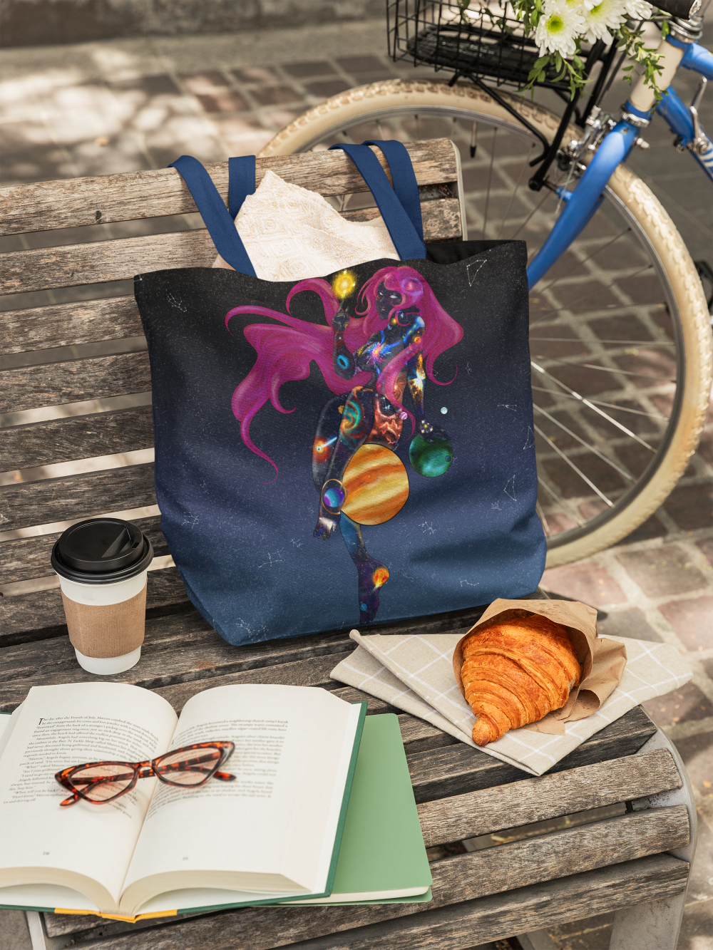 A wooden bench on a brick sidewalk holding a paper cup of coffee, an open book with glasses on it, and a bag with an galaxy-themed design. A croissant is on a paper towel nearby. A blue bike with a flower basket is in the background.