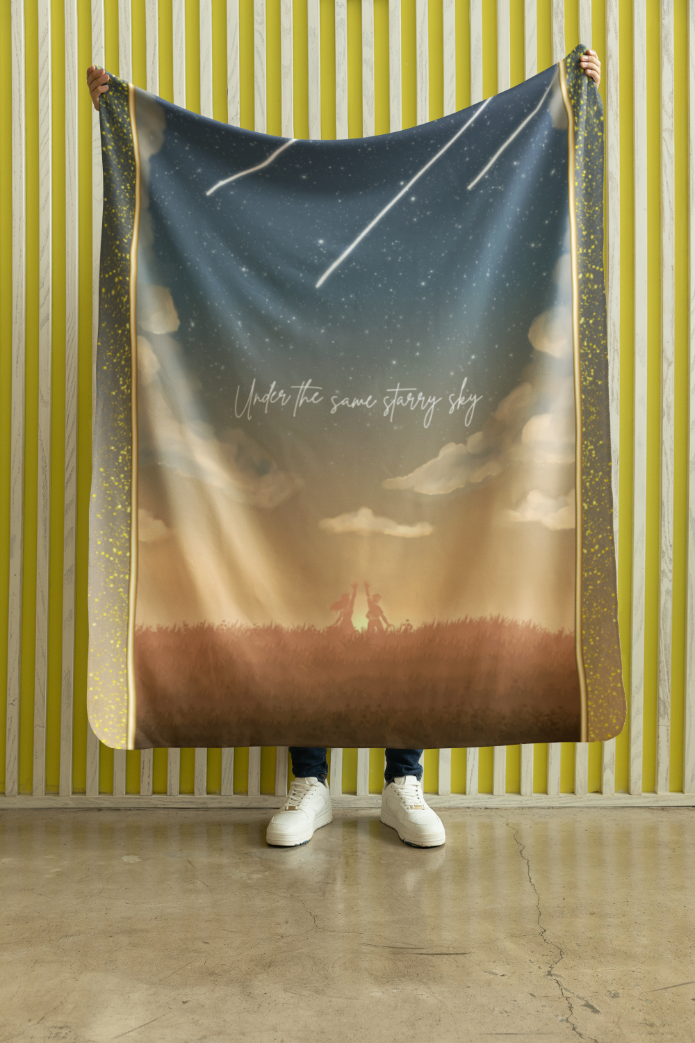 Person holding a blanket with a starry sky and sunset scene, the text "Under the same starry sky" written on it.