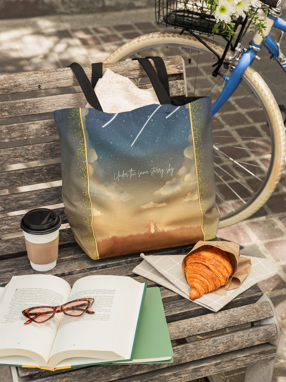 A wooden park bench with a tote bag, a coffee cup, an open book with glasses on it, a croissant on a napkin, and a bicycle with a basket of flowers nearby.