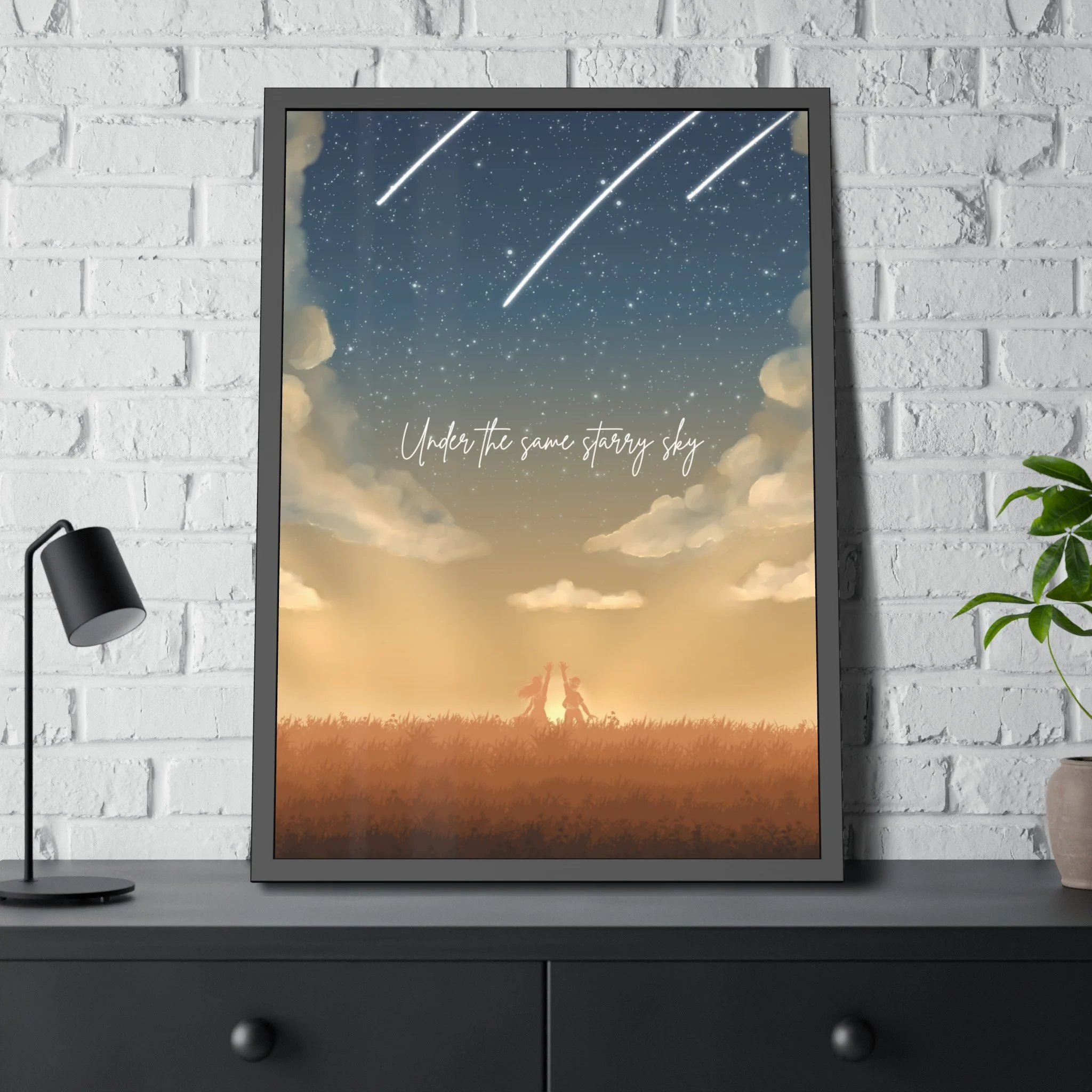 Framed poster on a desk showing a starry sky with shooting stars, a sunset with clouds, two children holding hands in a field, and the text 'Under the same starry sky'.