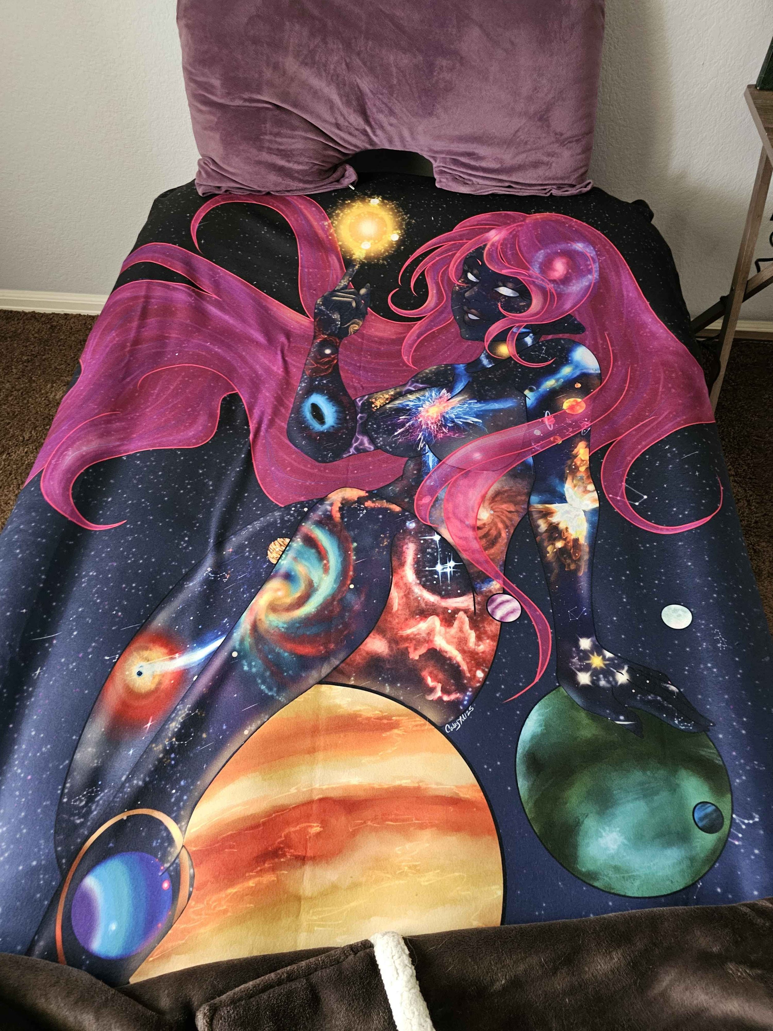 A blanket featuring an illustration of a cosmic-themed woman with pink hair, holding a glowing yellow orb, with stars and galaxies in the background.