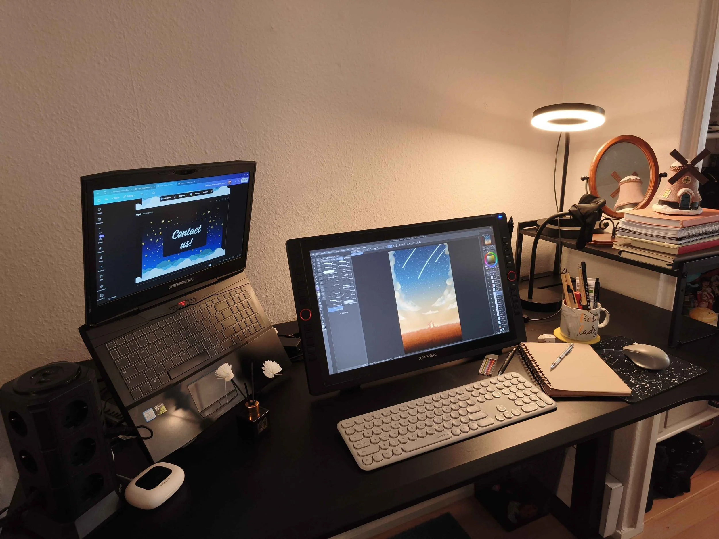 A cluttered black desk with two monitors, one displaying a "Contact us!" message and the other showing a digital artwork of a landscape with a meteor shower. The desk has a white keyboard, a mouse, a notebook, a mug filled with pens, and various small items. There are books, a lamp, and decorative objects on a small table behind the desk.