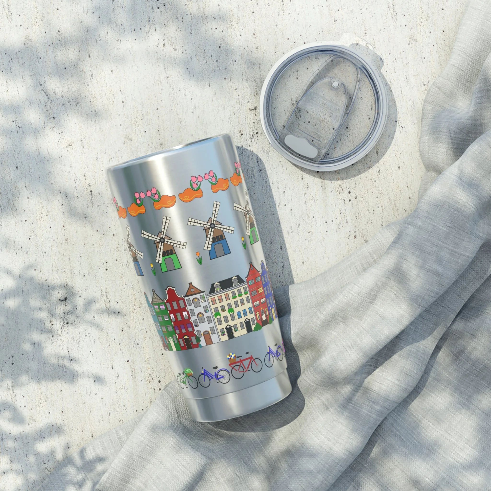 A stainless steel travel mug with colorful illustrations of windmills, tulips, houses, and bicycles on a light-colored surface, with the lid removed and placed nearby, and a light gray cloth underneath.