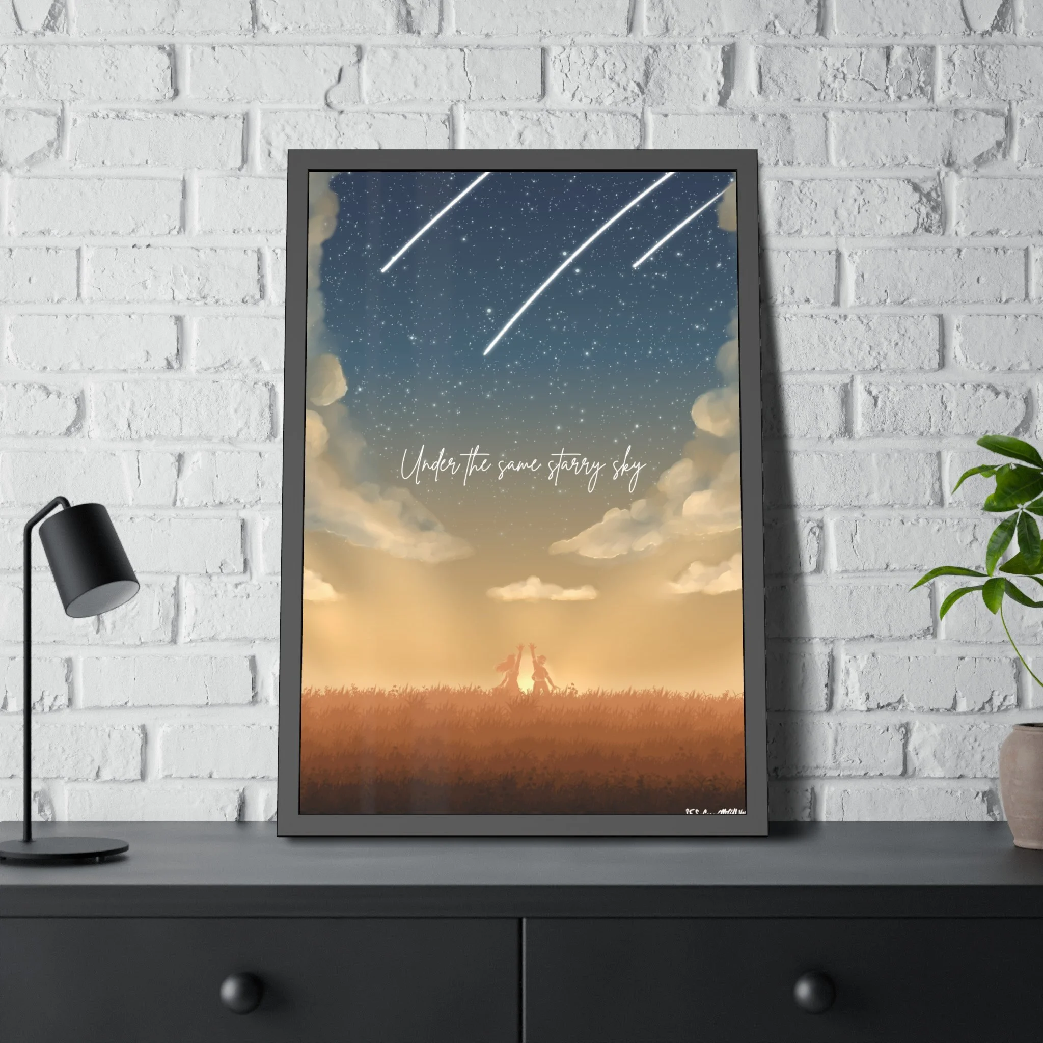A framed picture of a night sky with shooting stars and clouds, with the quote "Under the same starry sky" written on it, placed on a desk against a white brick wall, with a black desk lamp and a potted plant nearby.