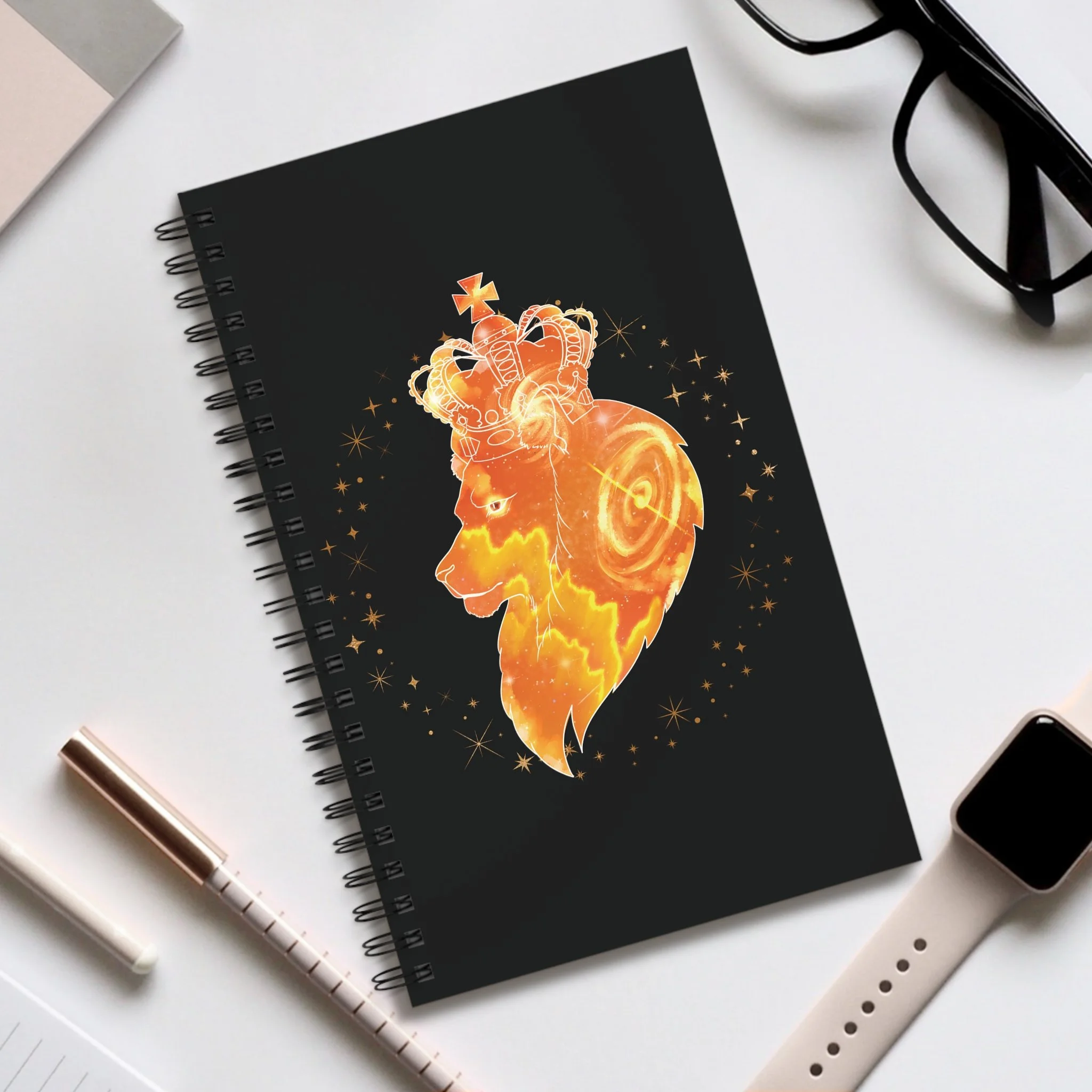 A black notebook with a glowing orange crown and dragon illustration on the cover, placed on a white desk with a pair of black glasses, a white and rose gold pen, and a white smartwatch around it.