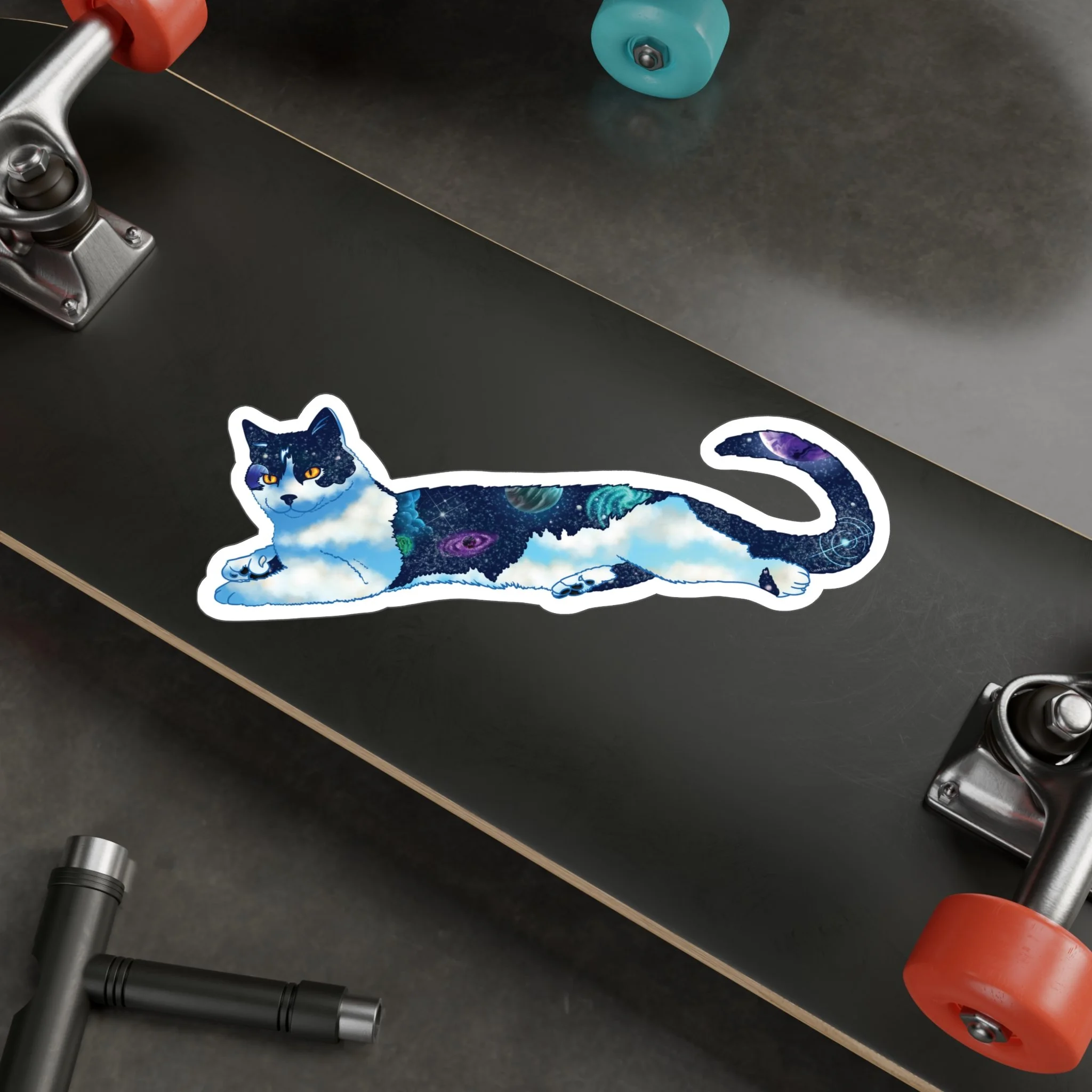 Close-up of a skateboard with cosmic-themed stickers, partially visible skateboard trucks, and skateboard wheels on a dark surface.