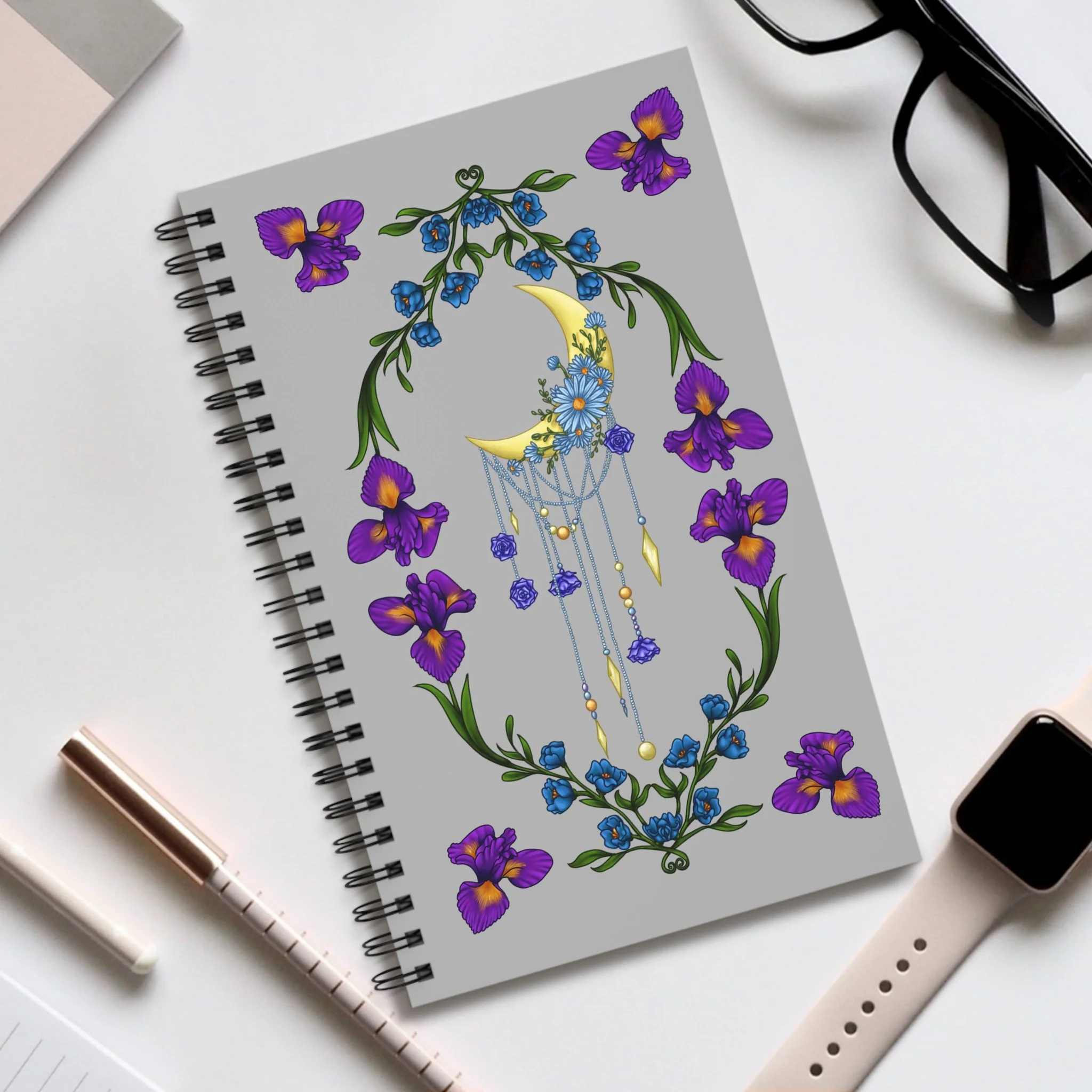 A notebook with a colorful design of flowers, a crescent moon, and a dreamcatcher with chains and beads on a white desk, accompanied by eyeglasses, a smartwatch, a pen, and notepads.