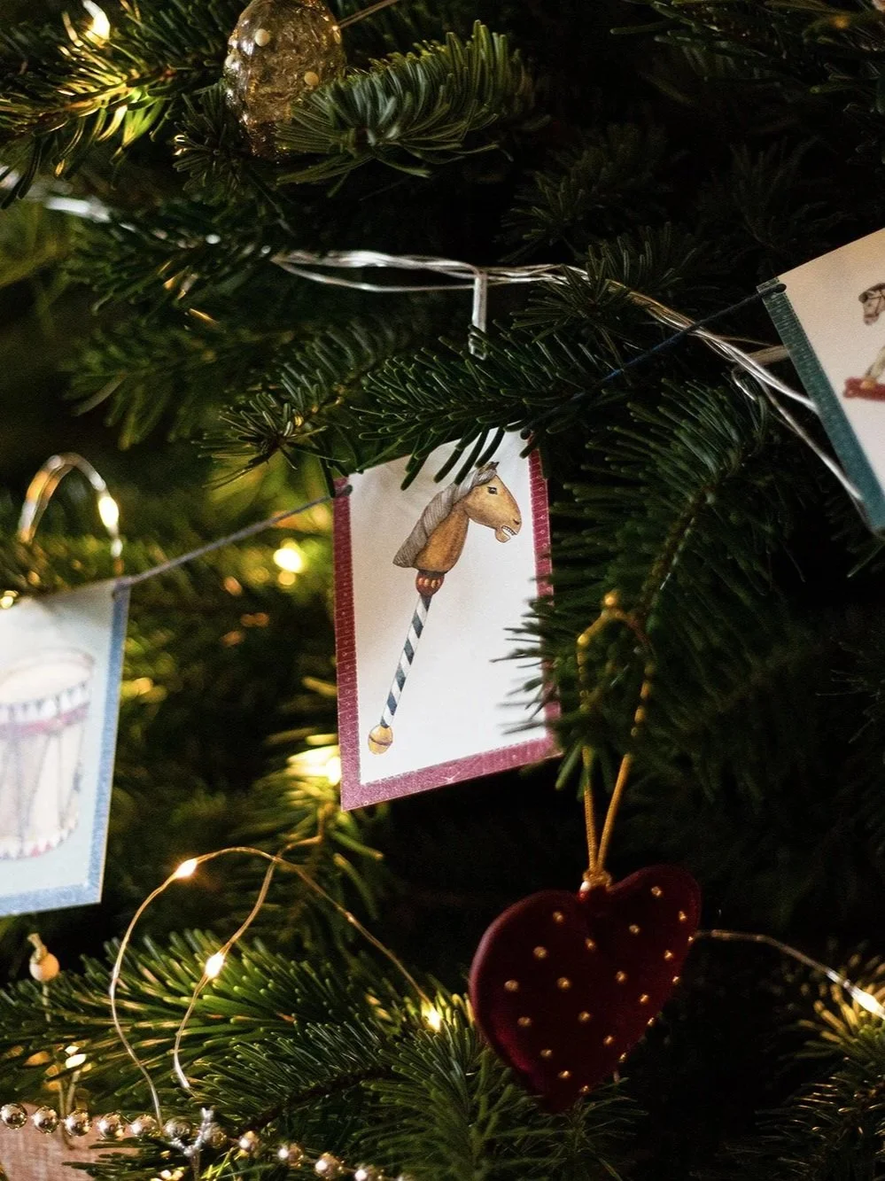 Decoración navideña en un árbol de Navidad con tarjetas ilustradas, como una del caballito de juguete y otra de una caja de música, además de un adorno en forma de corazón rojo