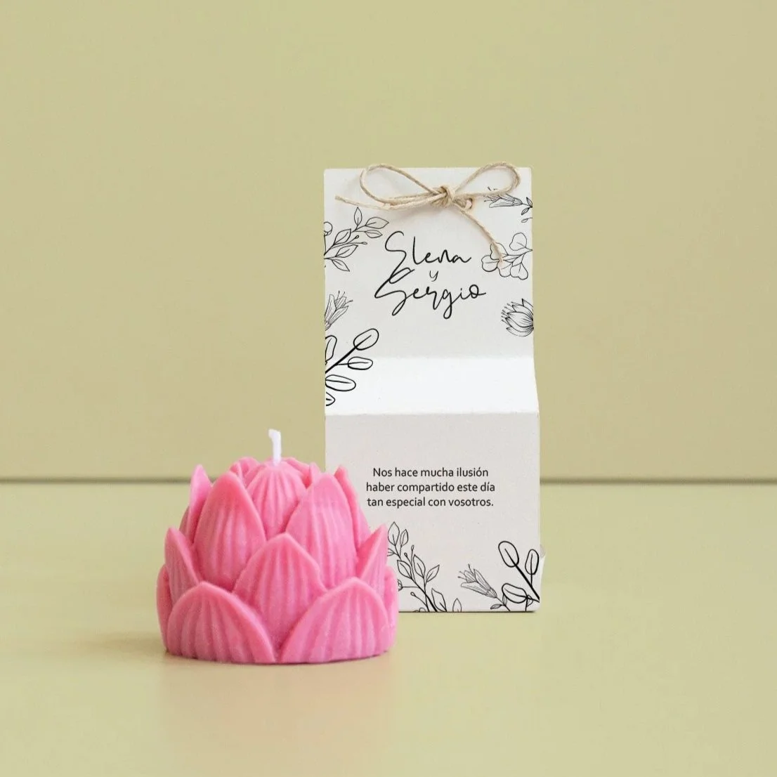 Una vela en forma de flor de color rosa y una tarjeta con nombres "Elena y Sergio" y mensaje en español, sobre fondo de mesa de color beige y pared beige claras.