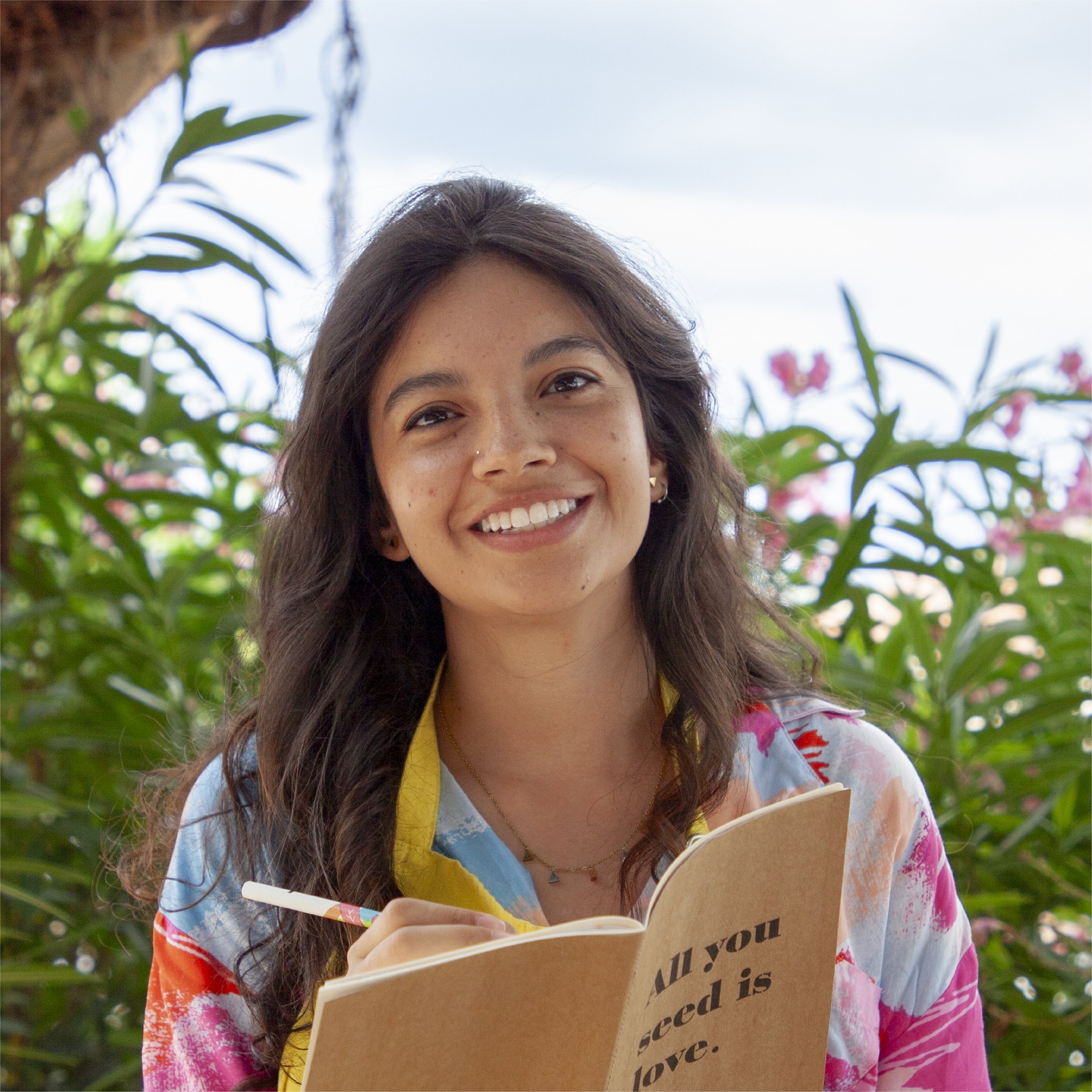 Mujer sonriendo leyendo un libro o cuaderno con fondo de plantas verdes y cielo ligeramente nublado.