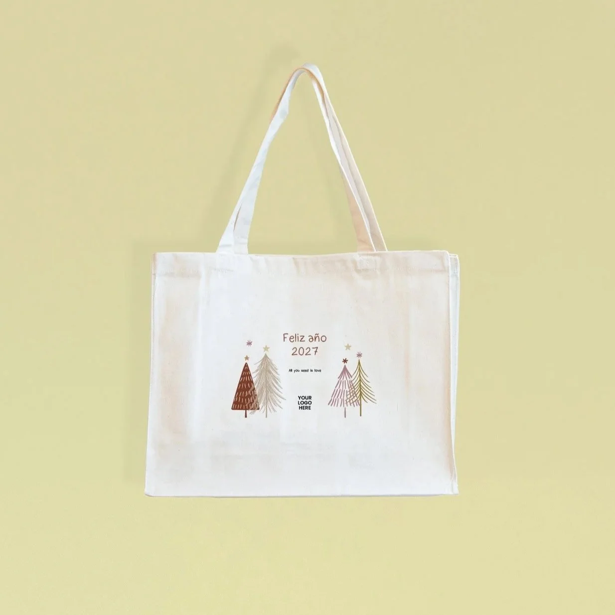 Bolsa de lona blanca con decoración navideña que incluye árboles y estrellas, y texto que dice 'Feliz año 2027' y 'All you need is love', además de un espacio para un logotipo personalizado