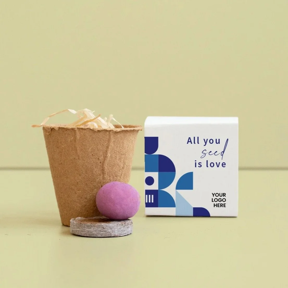 Un vaso de cartón con relleno de papel, una bola morada de semillas con una turba, junto a una caja blanca con diseño azul y texto en inglés que dice "All you seed is love" y espacio para logotipo.