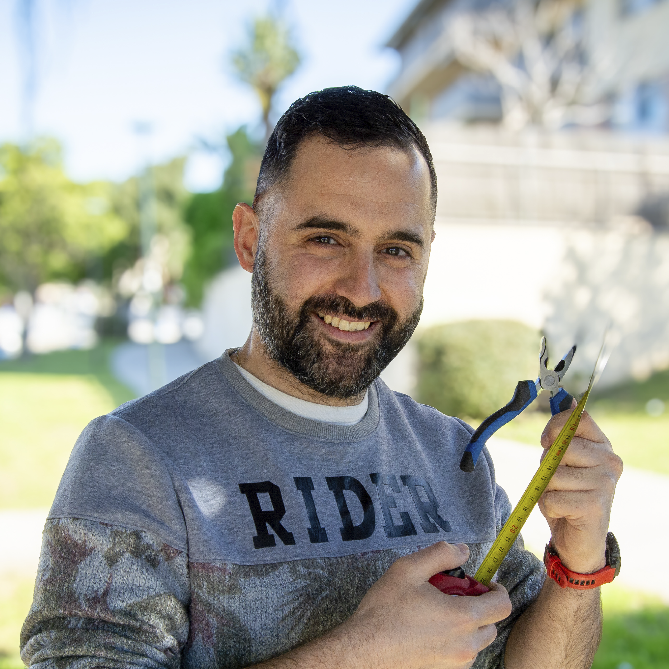 Un hombre con barba y cabello oscuro, sonriente, con una camiseta gris y un suéter de color azul oscuro, sosteniendo una cinta métrica amarilla en el exterior en un día soleado con fondo de árboles y cielo azul.