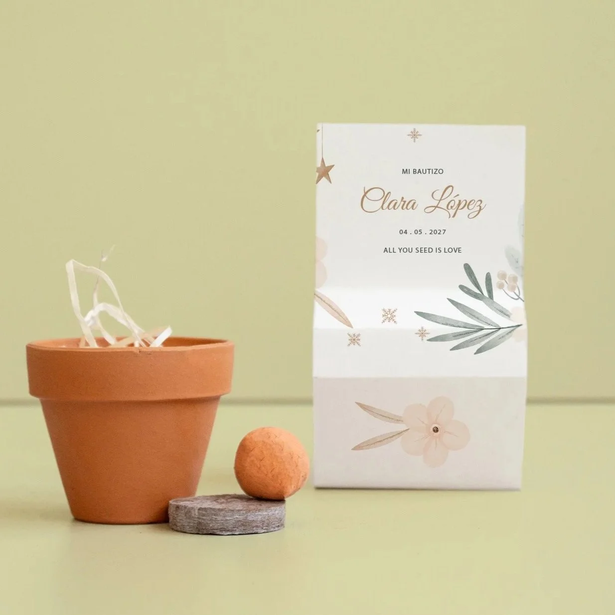 Una maceta de barro con objetos blancos dentro, una bola de color terracota sobre un pedal de madera, y una tarjeta de bautizo con diseño floral y texto en español, en un fondo de color verde claro.